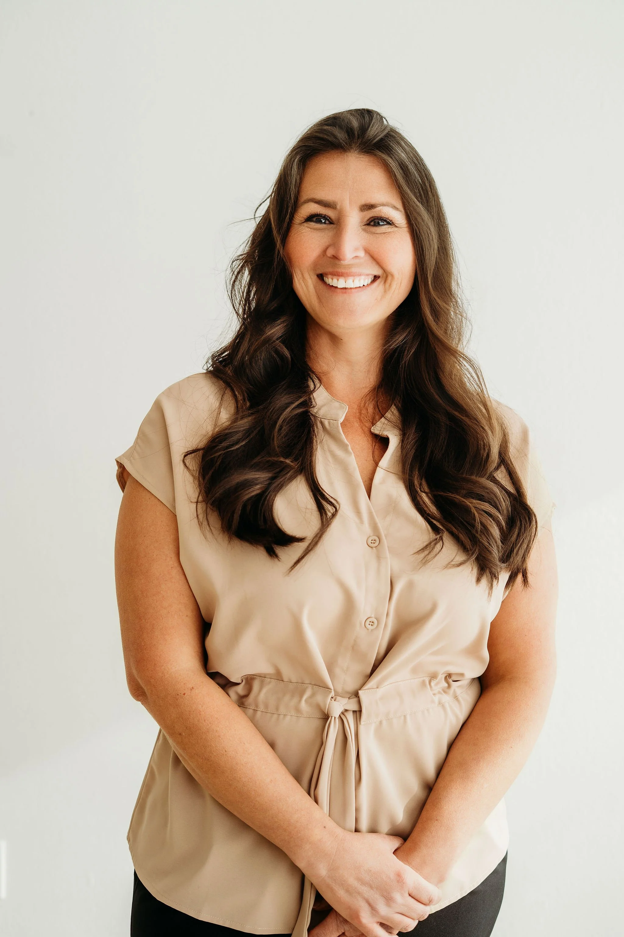 A woman smiling, with long brown hair, sitting on a brown leather chair against a beige wall.