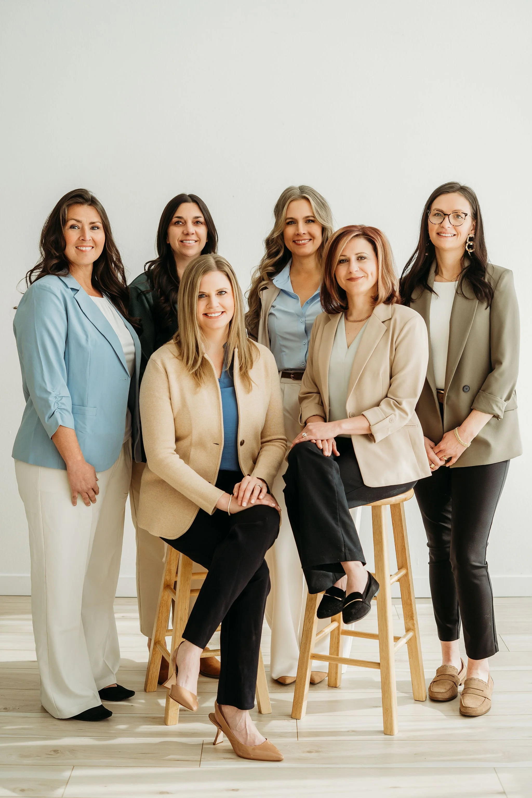 Six women in business attire posing together in front of a plain wall, some sitting on stools and some standing, smiling at the camera.