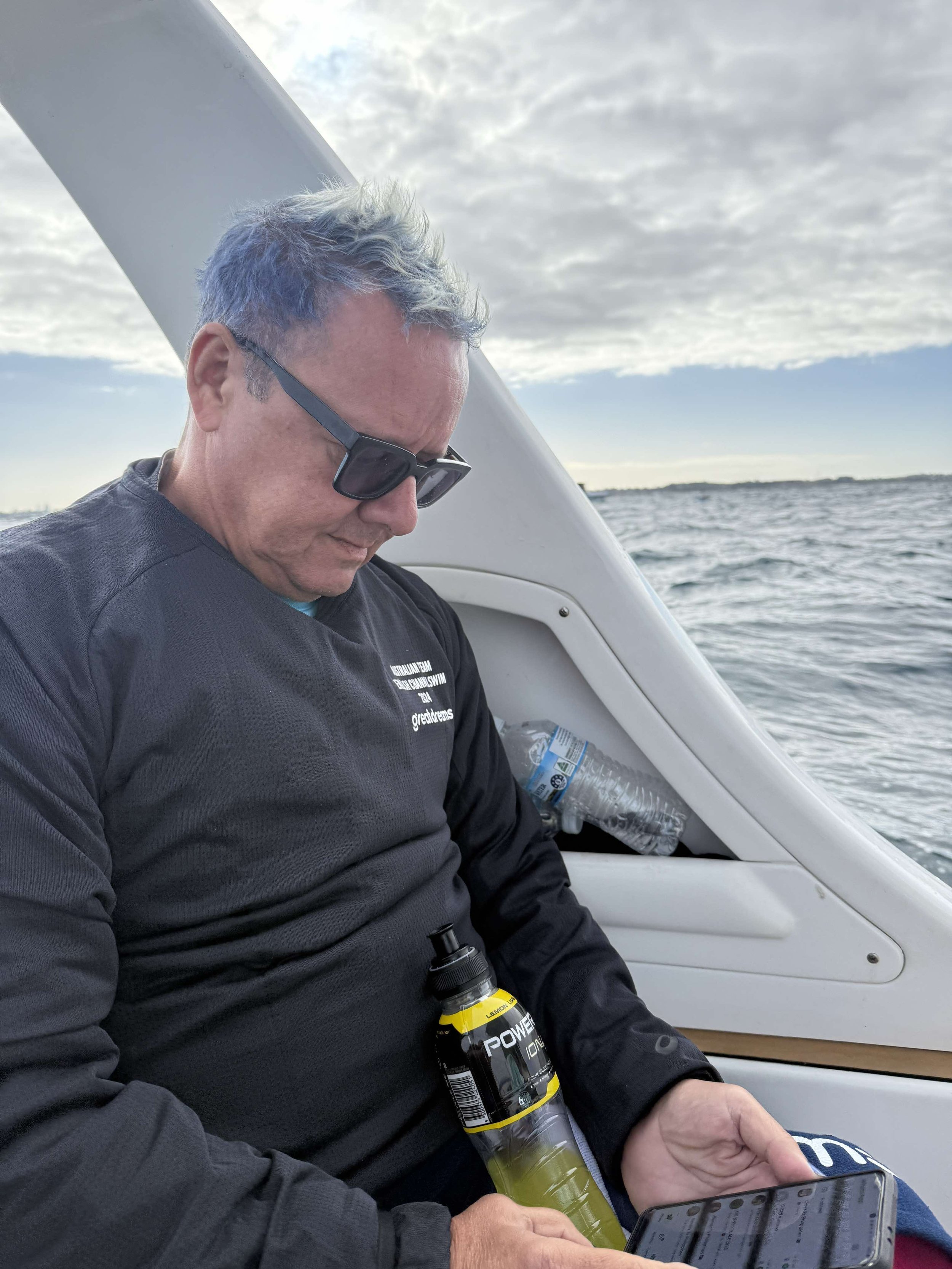 Guide swimmer Phil sitting on the support boat the horizon behind him, getting updates on the conditions from a tablet on his lap. A bottle of yellow gatorade also in close reach.
