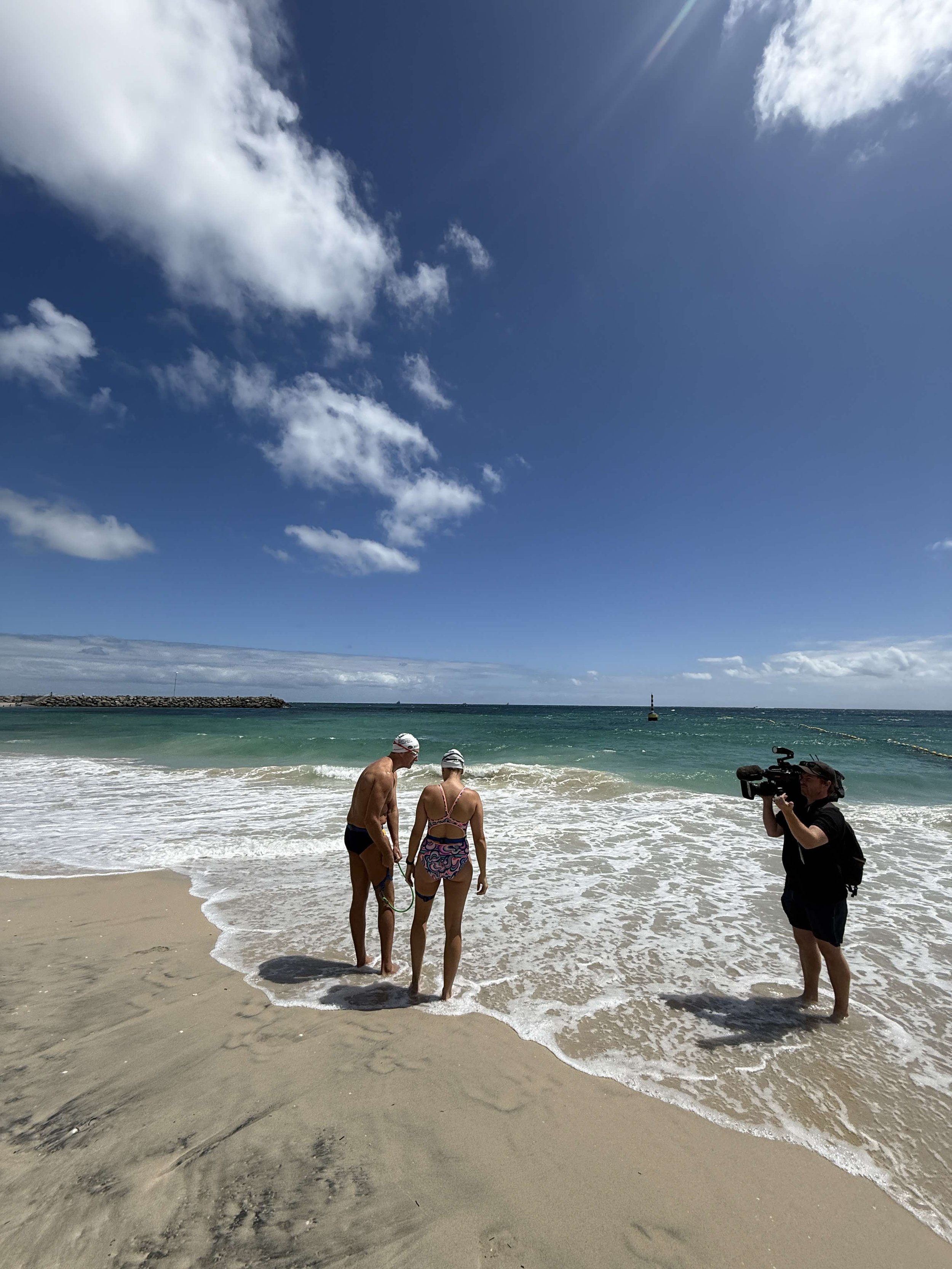 Gerrard and Kristy standing on the beach looking out over the water. A TV cameraman is standing on the right of the short recording them