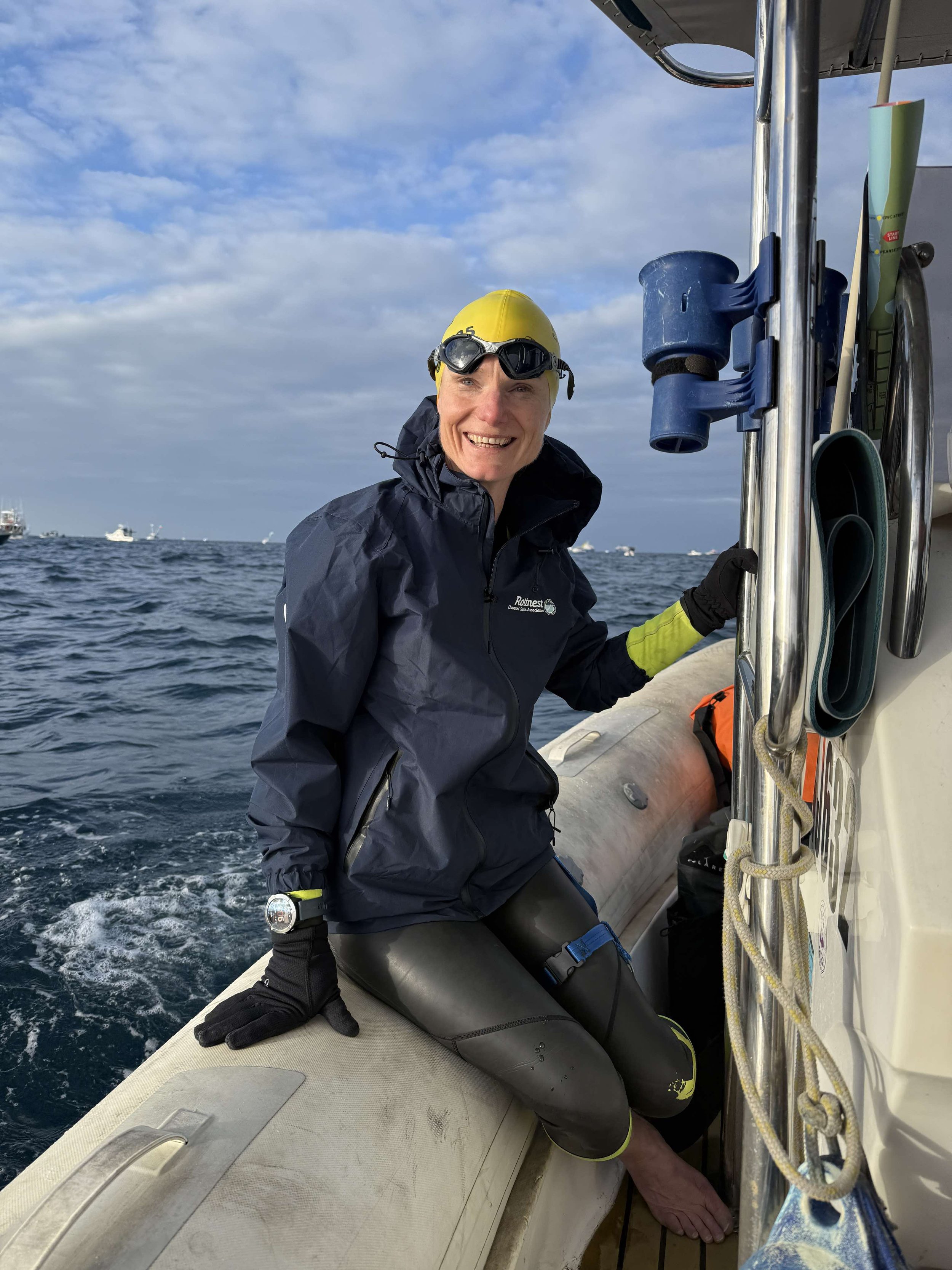 Guide swimmer Jo keeping watch from the support boat in a black wetsuit, yellow cap and goggles. The ocean and the sky behind her.