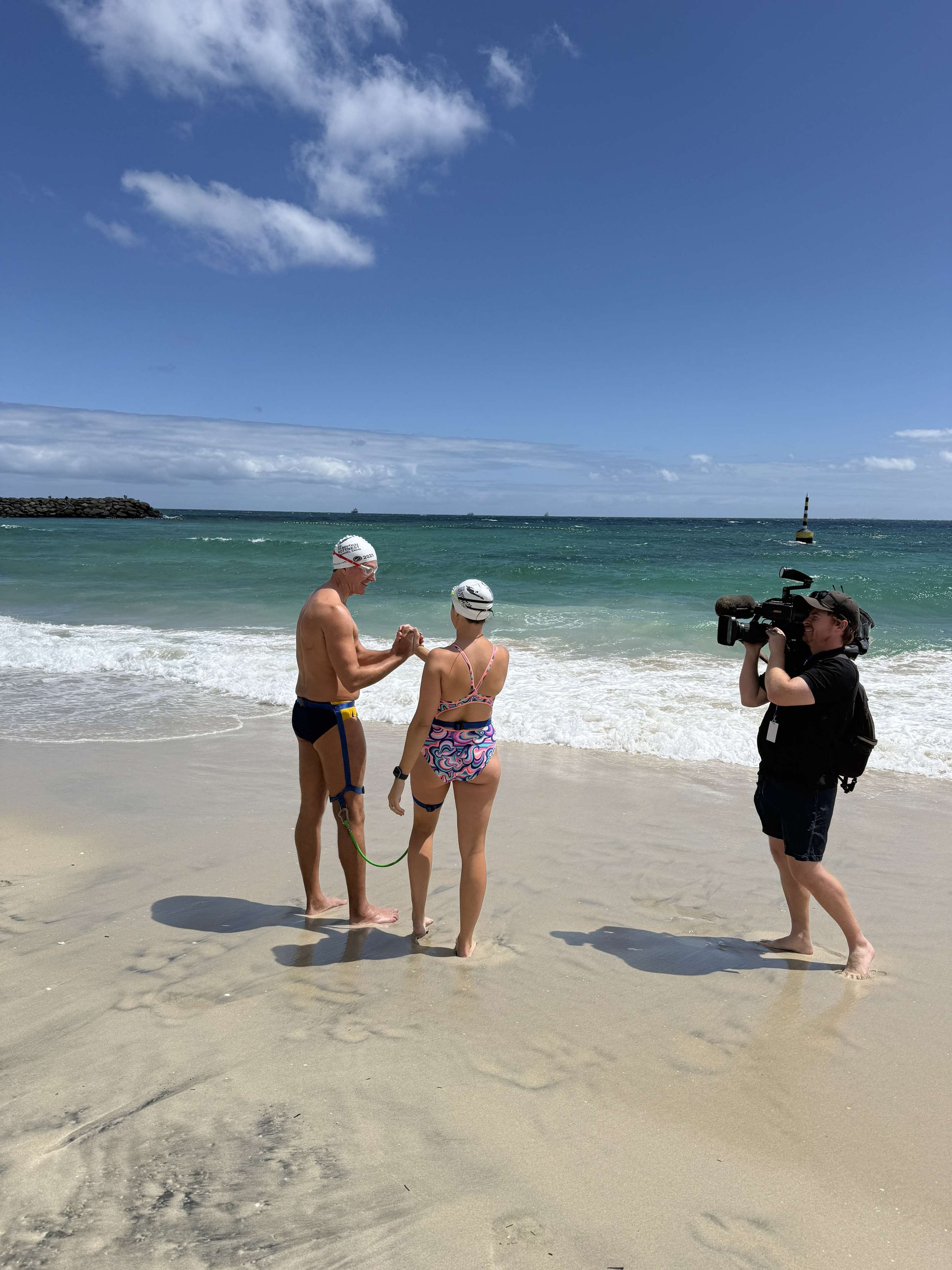 Gerrard and Kristy on the beach about to enter the water. They are shaking hands, and both are wearing swim tethers. A cameraman from Channel 10 stands in the right recording them.