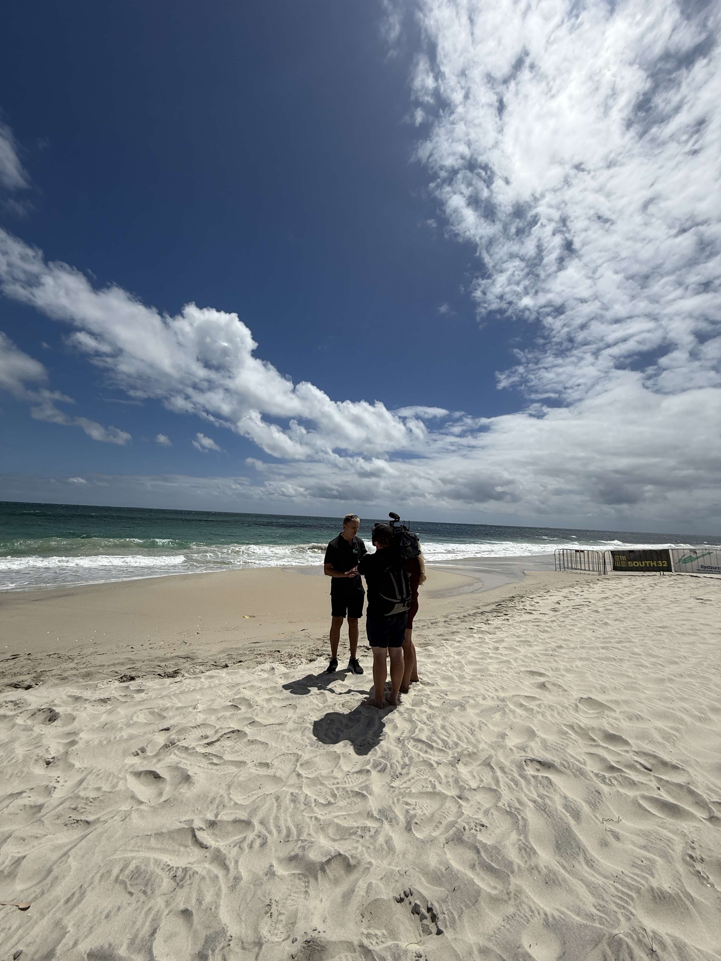 Wide shot of Gerrard being interviewed by Channel 10 on the beach prior to the swim. White sand in the bottom half, scattered clouds and blue sky across the top.