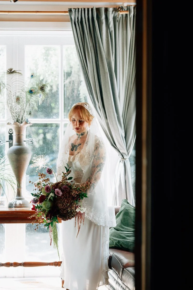 A bride in a white dress with tattoos on her arms and neck, holding a large bouquet of flowers, standing near a window with gray curtains and sunlight streaming in, inside a cozy room with a vase and decorative plants.