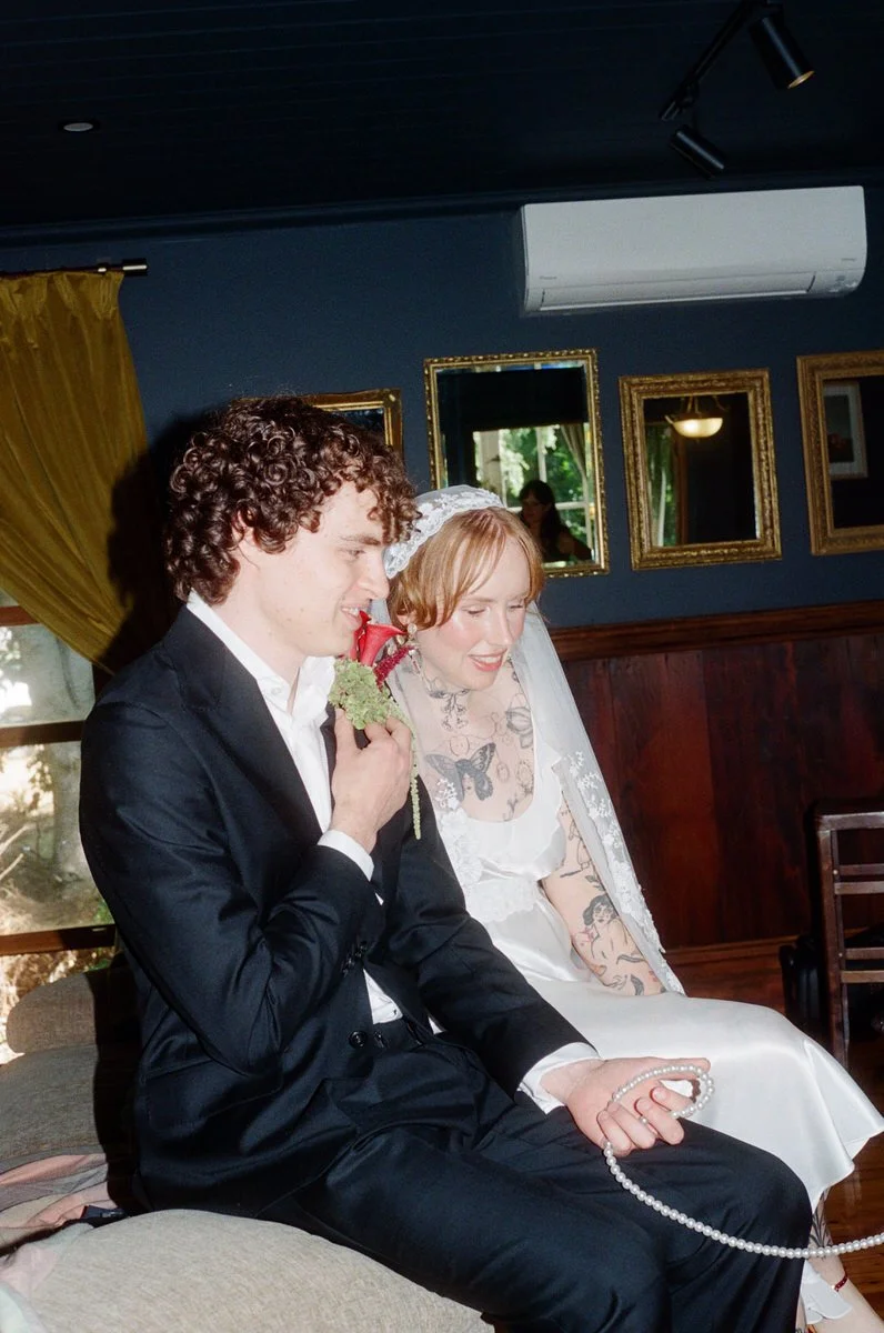 A bride with tattoos and a groom in a black suit sitting together in a room with dark blue walls, framed pictures, and gold curtains, smiling and holding wedding accessories.