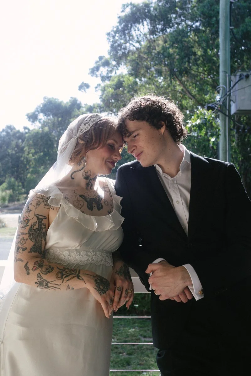 A bride and groom with their foreheads touching, outdoors on a sunny day, with trees and a utility pole in the background.