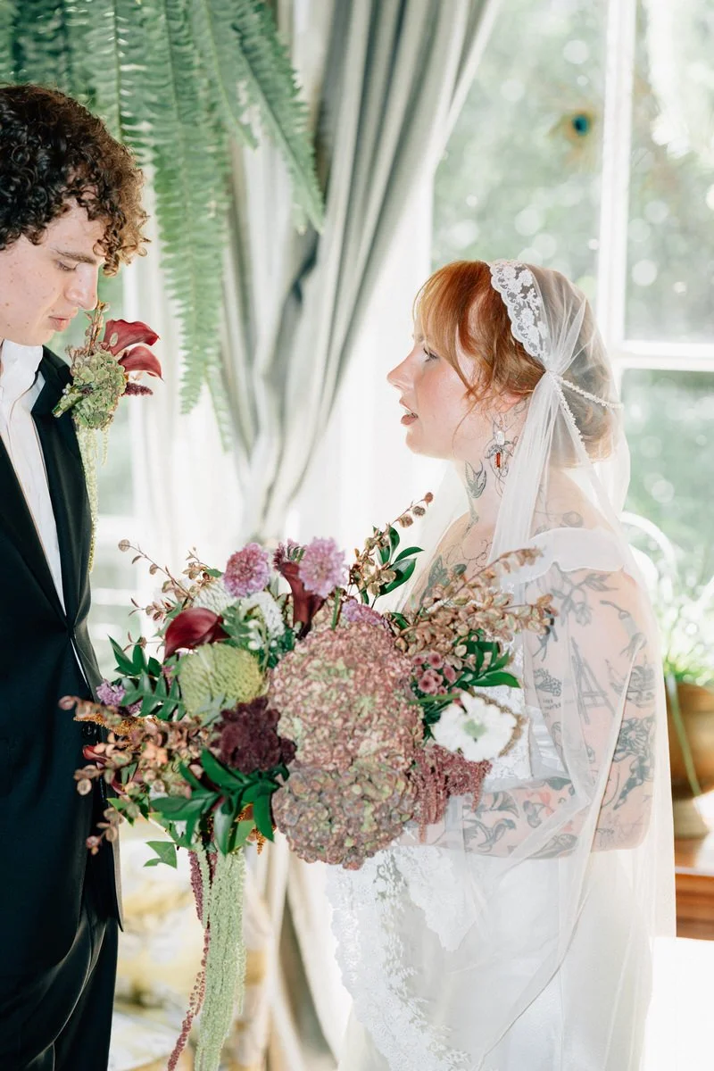 A bride holding a large bouquet of flowers looking at her groom during a wedding ceremony indoors near a window with curtains.