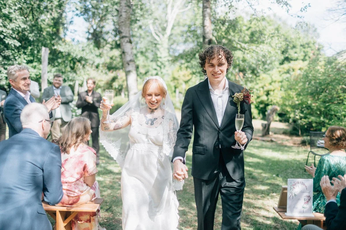 A newlywed couple holding hands and smiling, walking outdoors during a wedding celebration, with guests clapping and enjoying the event in a lush, green park.