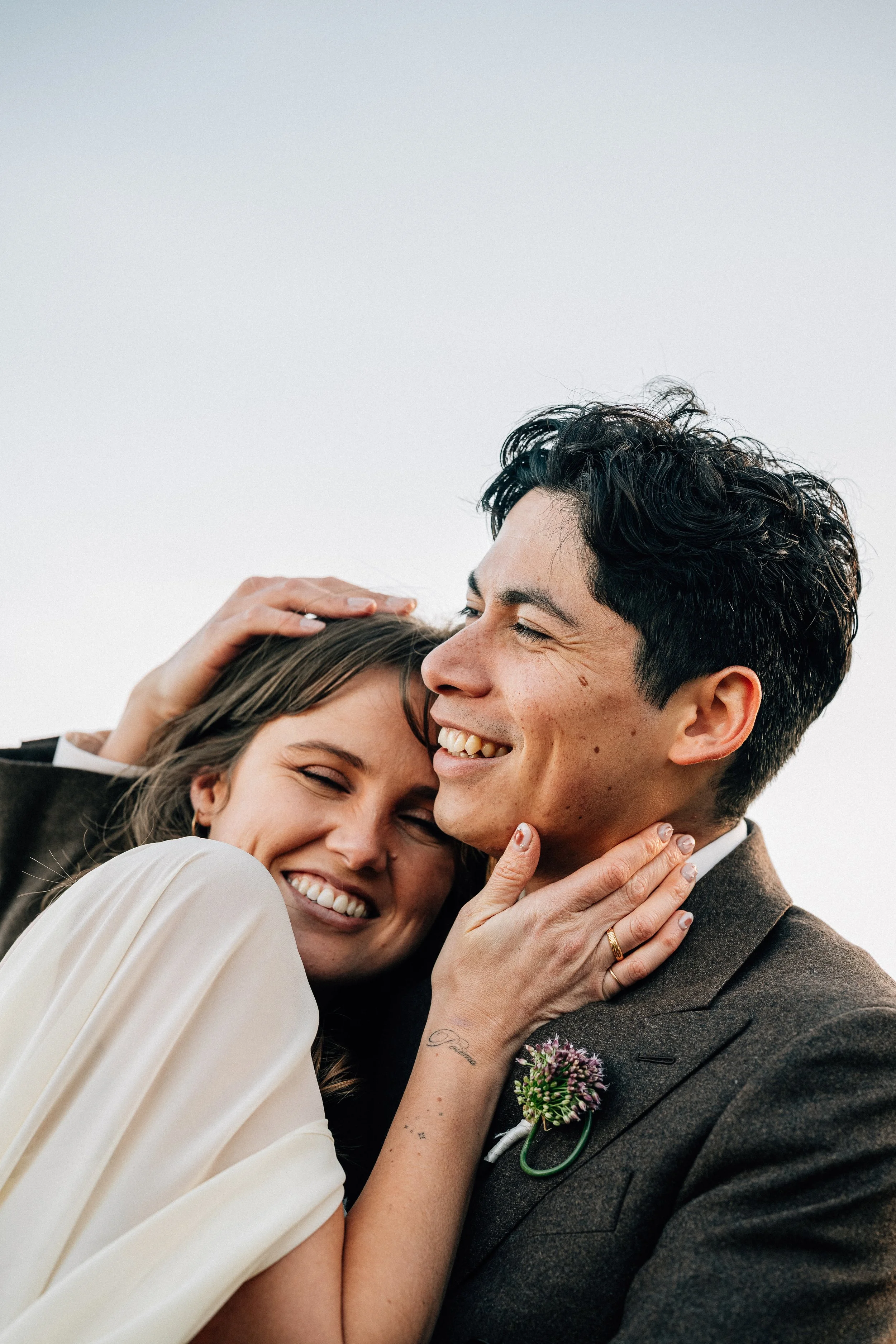 A happy couple hugging each other and smiling, with the woman resting her head on the man's shoulder. The man has dark, curly hair and is wearing a suit with a boutonniere, and the woman has light brown hair and is wearing a cream dress. The woman's hand is gently touching the man's face, and they appear to be at an outdoor event with a clear sky.