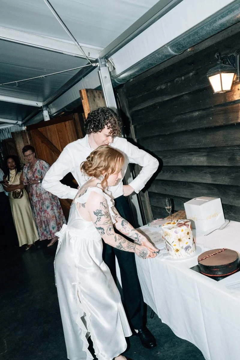 Two women cutting a wedding cake at a celebration indoors, with guests in the background.