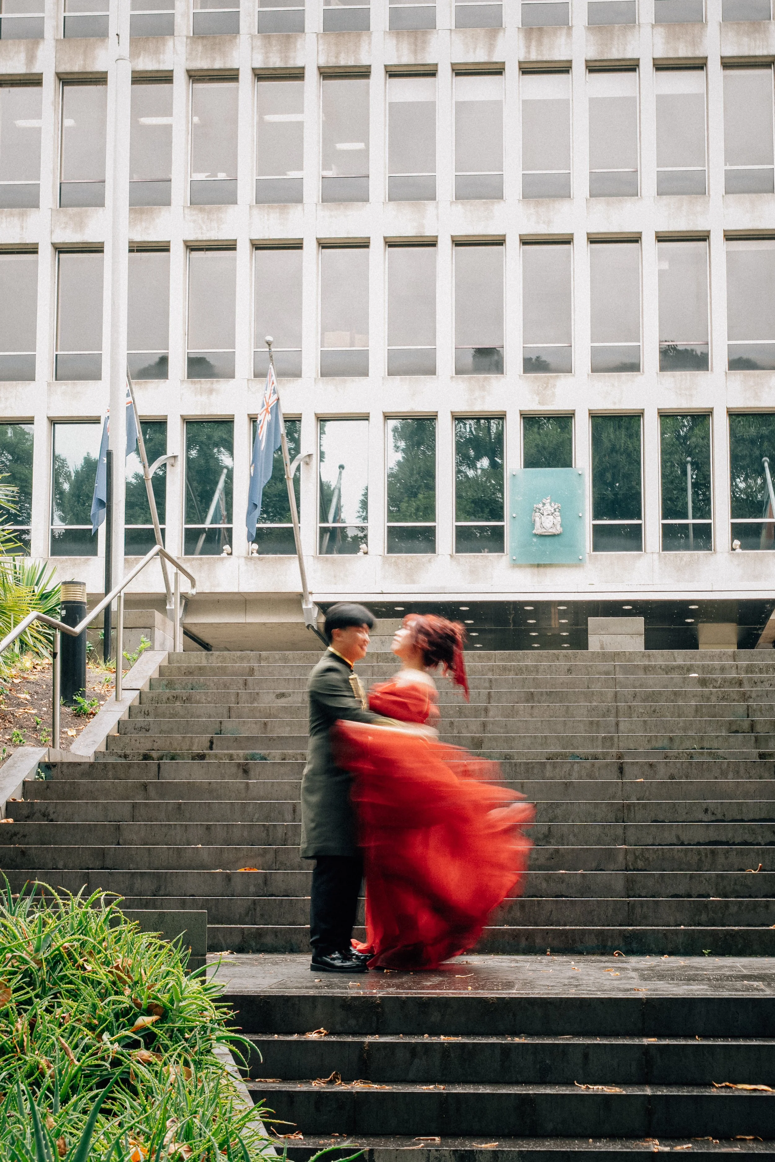 Couple dancing on front steps of government building with flags and coat of arms, greenery in foreground, blurred motion of woman in red dress.
