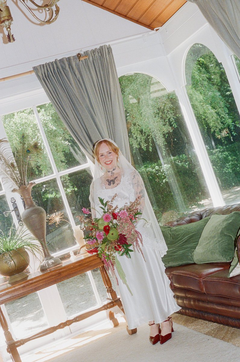 A bride standing next to a wooden table with a large floral bouquet, in a sunlit room with green trees outside the window, a tufted leather sofa with green cushions, and decorative vases on the table.