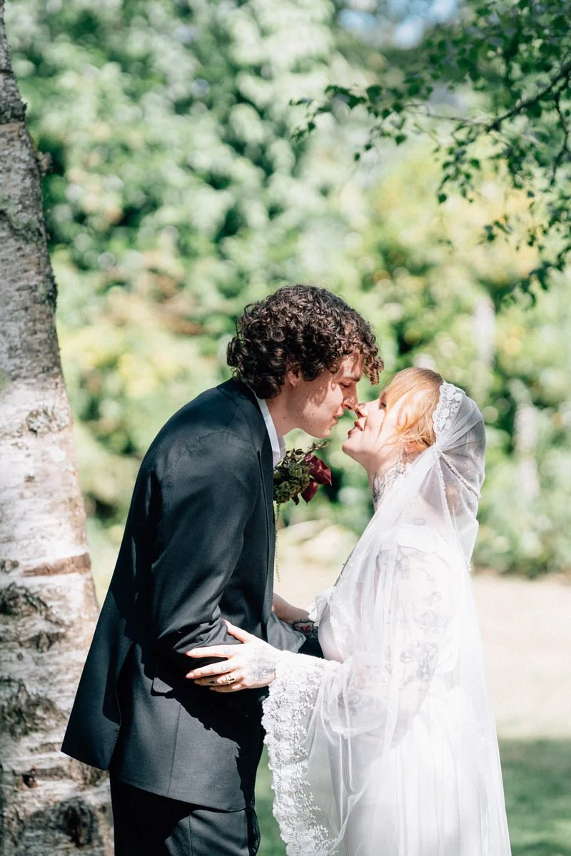 A bride and groom are outdoors, sharing a kiss underneath a tree on their wedding day. The bride is wearing a white dress with lace details and a veil, and the groom is dressed in a black suit with a boutonniere.