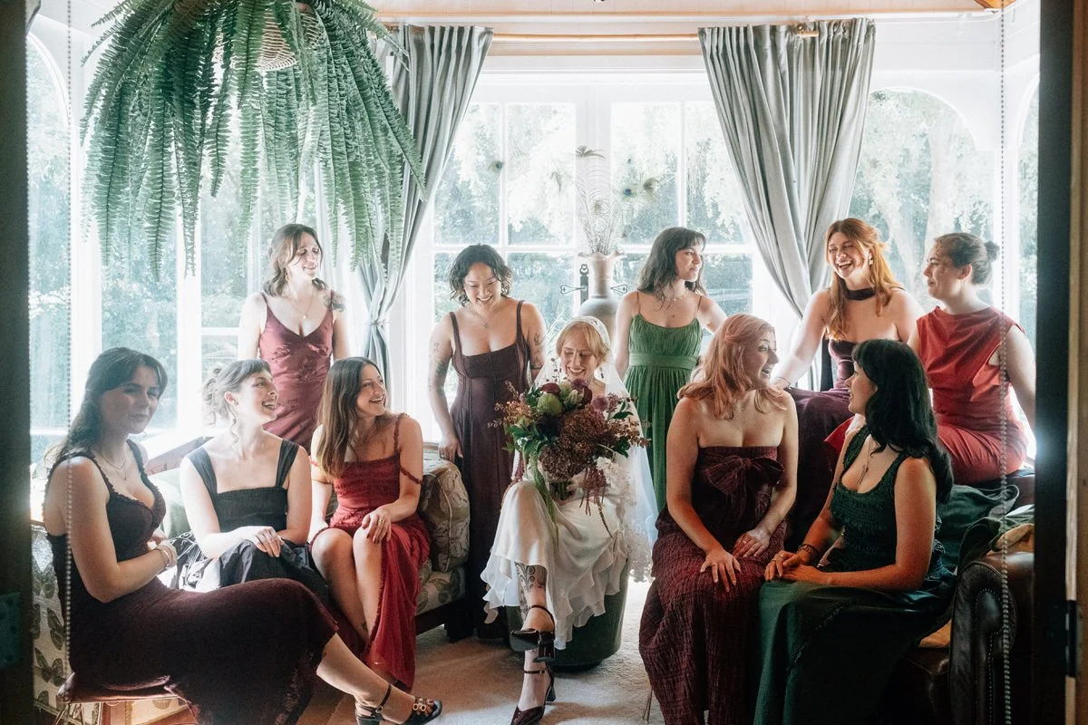 A group of women gathered indoors near large windows, with some sitting and others standing, in elegant dresses, celebrating a special occasion with a woman in a white dress holding a bouquet of flowers.