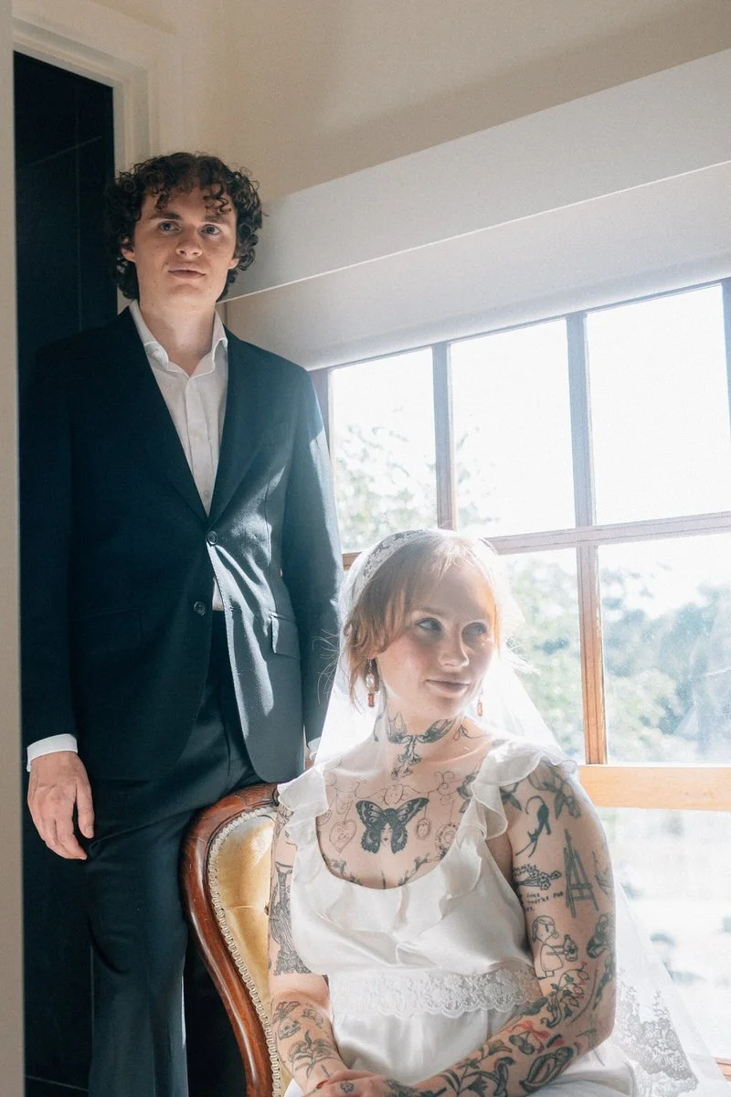 A young woman with tattoos and a vintage dress sitting near a large window, and a young man in a suit standing behind her.