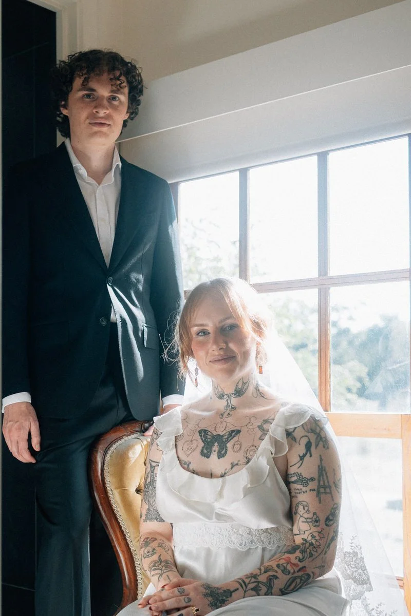 A couple dressed in wedding attire standing near large windows with trees outside, with the bride wearing a lace veil and the groom in a dark suit, holding hands and smiling.