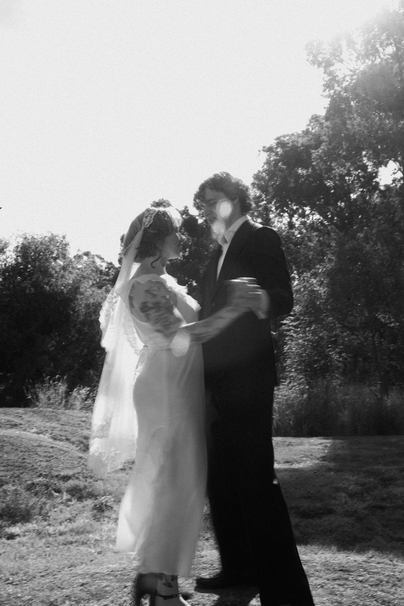 A couple in wedding attire sharing a romantic moment outdoors, holding each other, with trees in the background, in black and white.