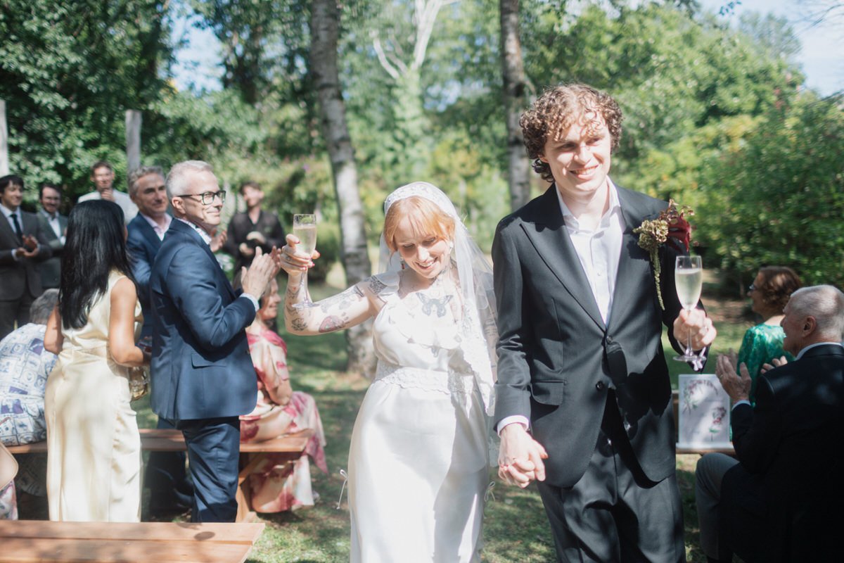 A newlywed couple holding hands and drinking champagne at an outdoor wedding reception. The bride has blonde hair, tattoos, and a white dress. The groom has curly hair, wears a black suit, and has a boutonniere. Guests are clapping and smiling in the