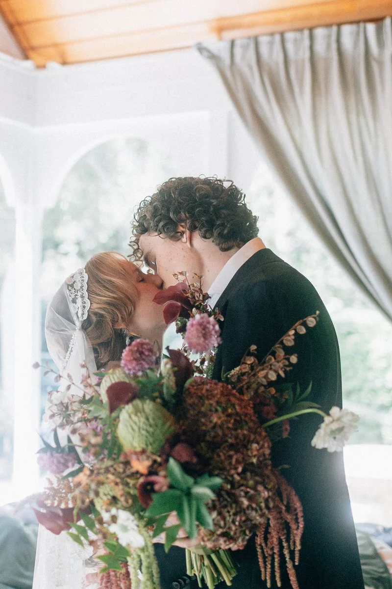 A bride and groom share a kiss indoors, with the bride holding a large bouquet of pink, purple, white, and green flowers. The bride wears a veil and wedding dress, and the groom is dressed in a dark suit and white shirt.