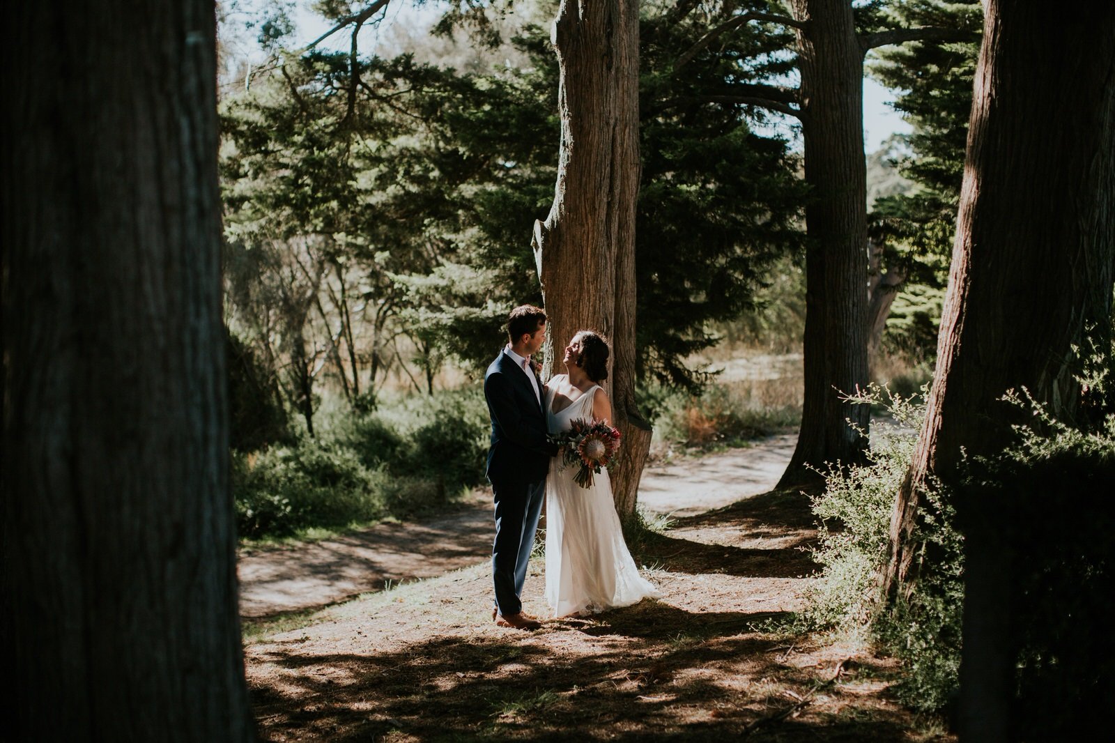 Bride and groom standing in a wooded area surrounded by trees, with sunlight filtering through the branches. The bride holds a bouquet of flowers.