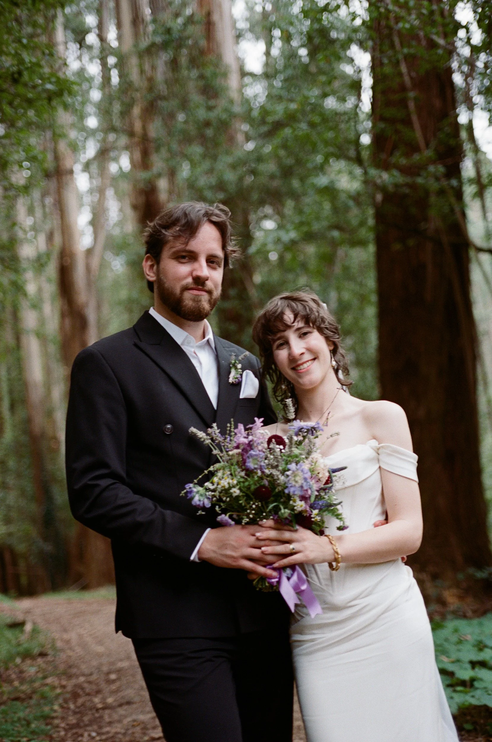 A couple in wedding attire posing in a forest, the groom in a black suit and the bride in a white dress holding a colorful bouquet.