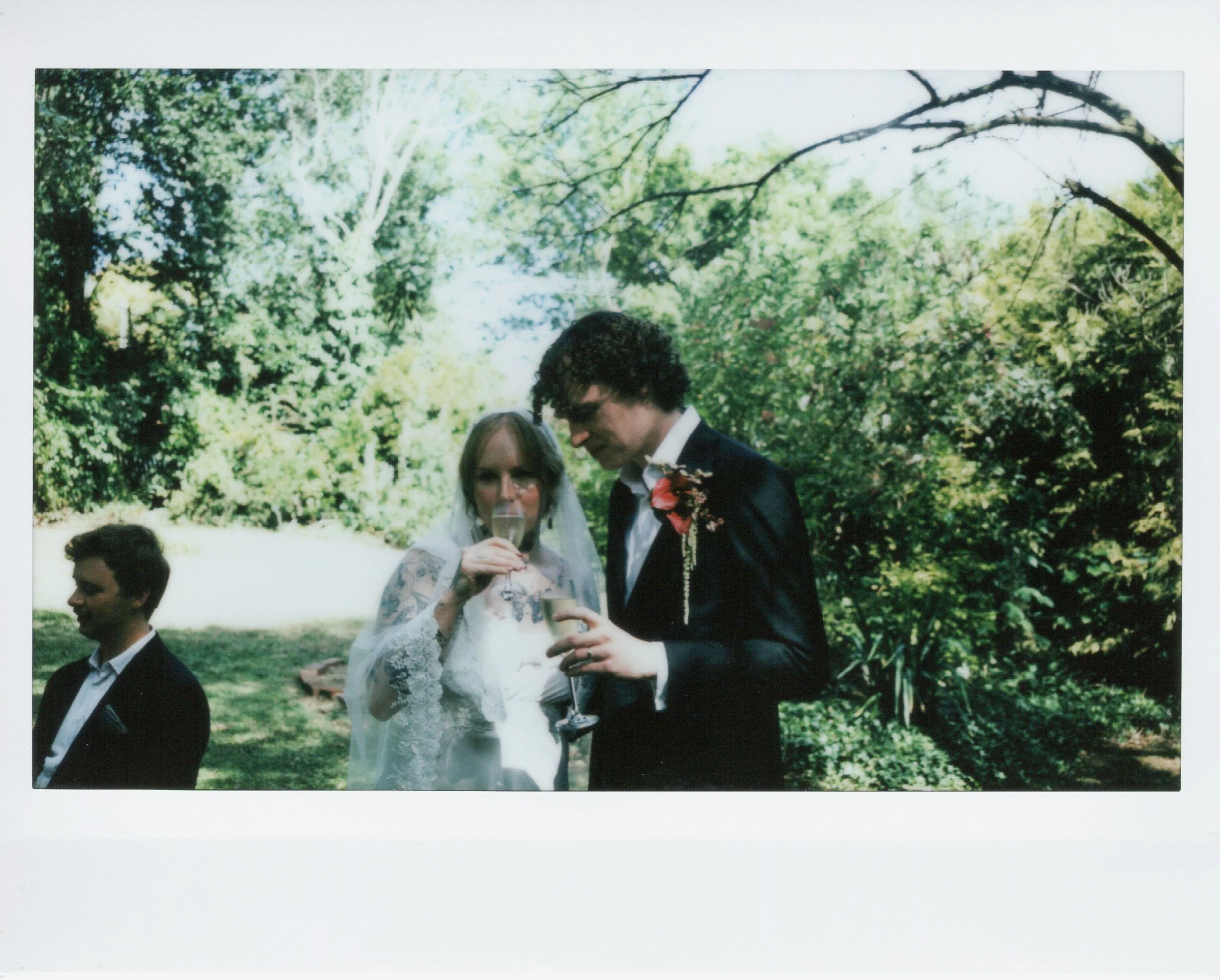 A bride and groom at an outdoor wedding, the bride drinking from a glass and the groom holding a drink, with a man sitting nearby under trees.