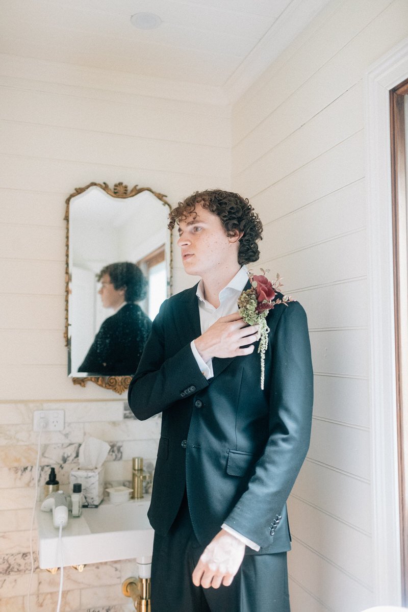 A young man in a black suit and white shirt stands in a room with beige paneling, holding a small boutonniere with red and green flowers near his chest. There is a mirror behind him reflection showing his profile, and a bathroom sink with toiletries 