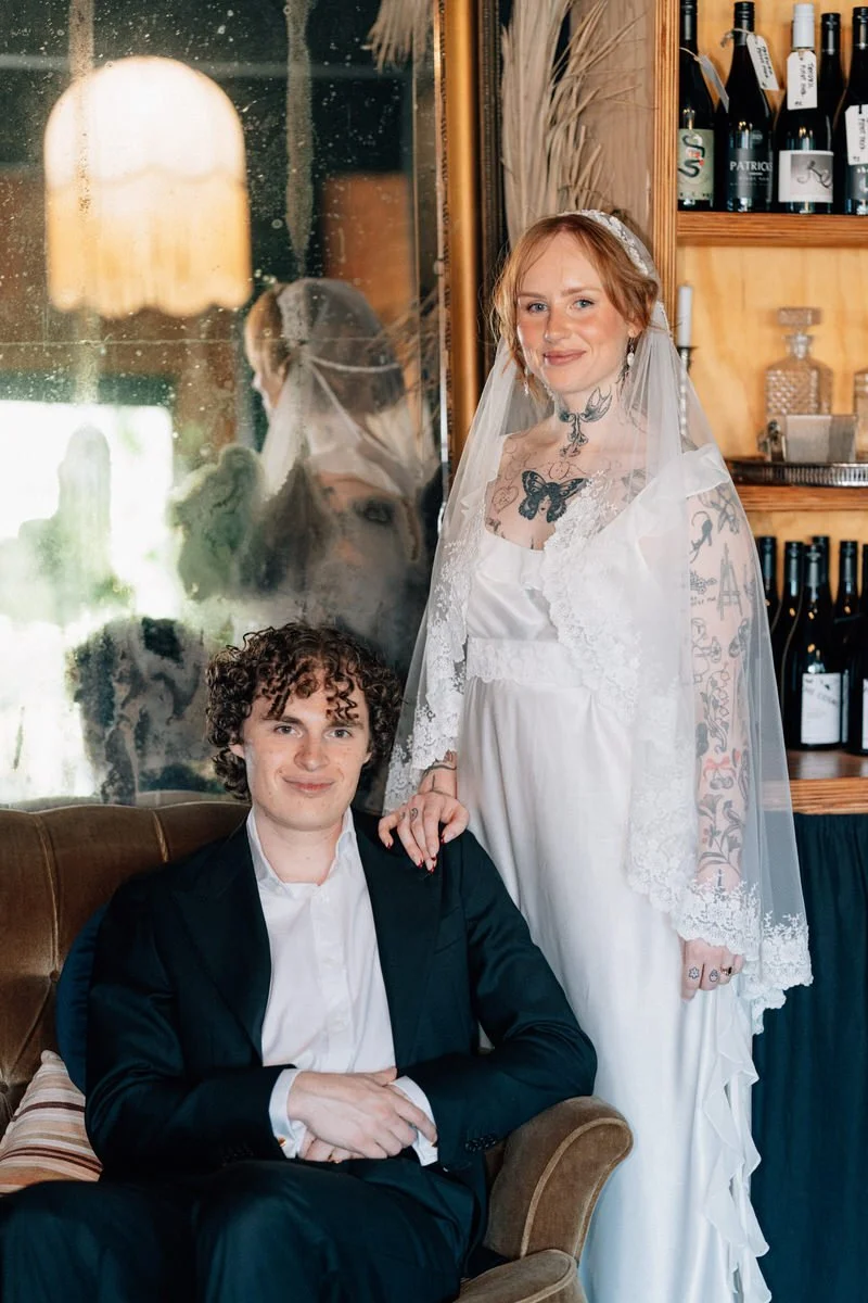 A bride in a white wedding dress and veil standing next to a groom in a black suit sitting on a brown chair in a cozy indoor setting with a wooden shelf of wine bottles in the background.