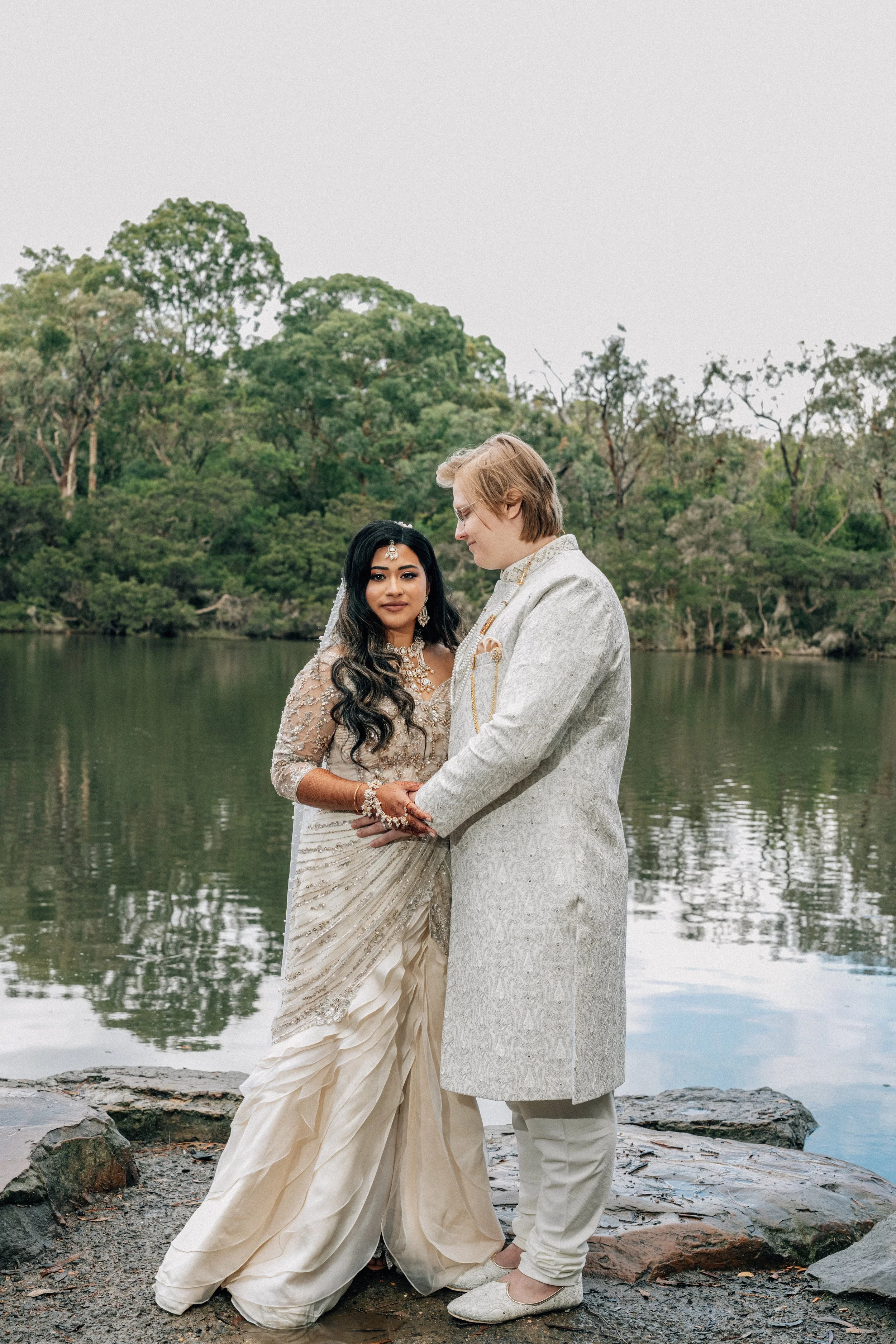 A bride and groom stand close together by a lake, holding hands, with trees and water in the background, during a wedding ceremony.