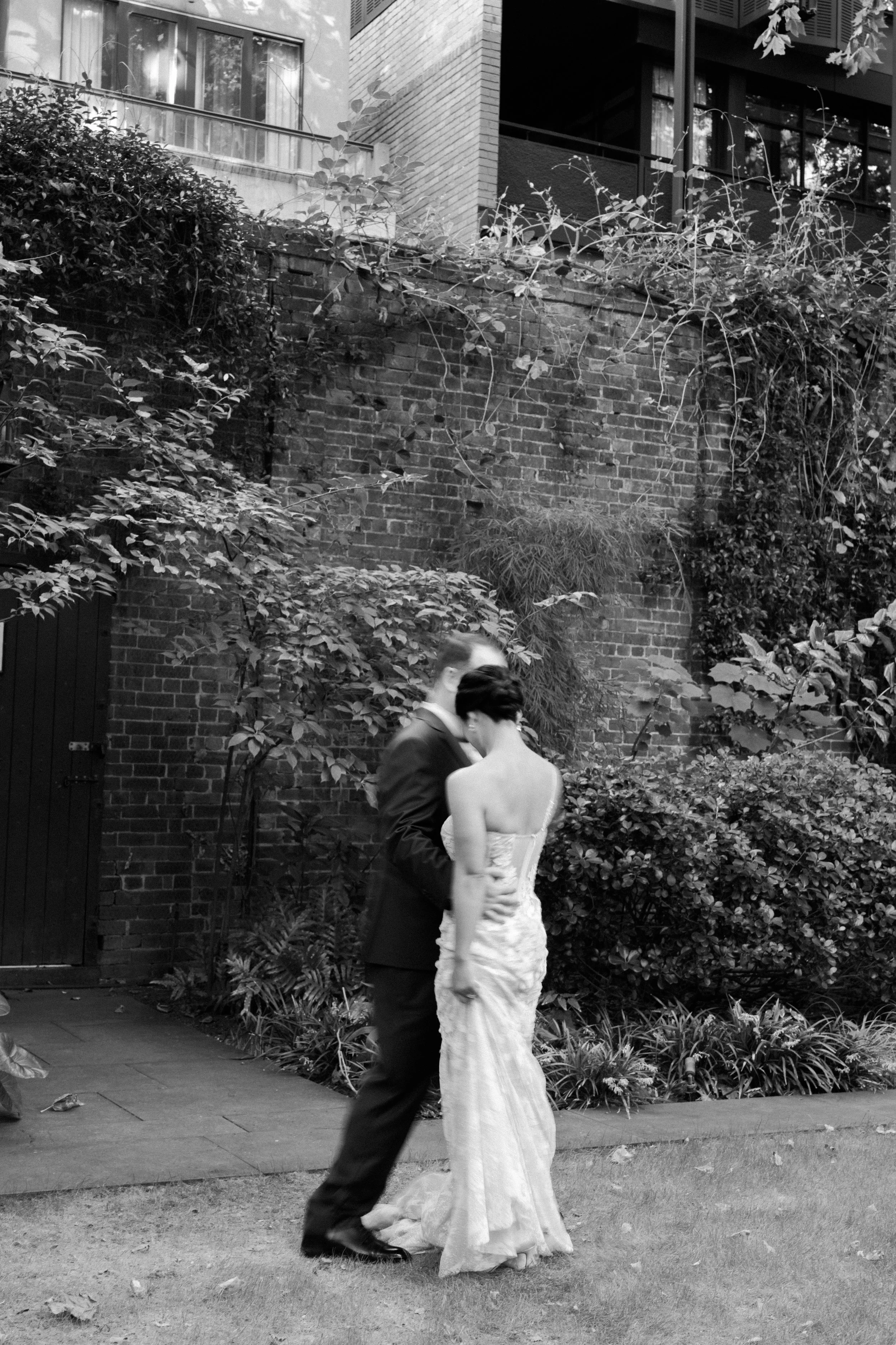 A black and white photo of a couple dancing outdoors in front of a brick wall and lush greenery. The man is wearing a suit and the woman is in a strapless gown.
