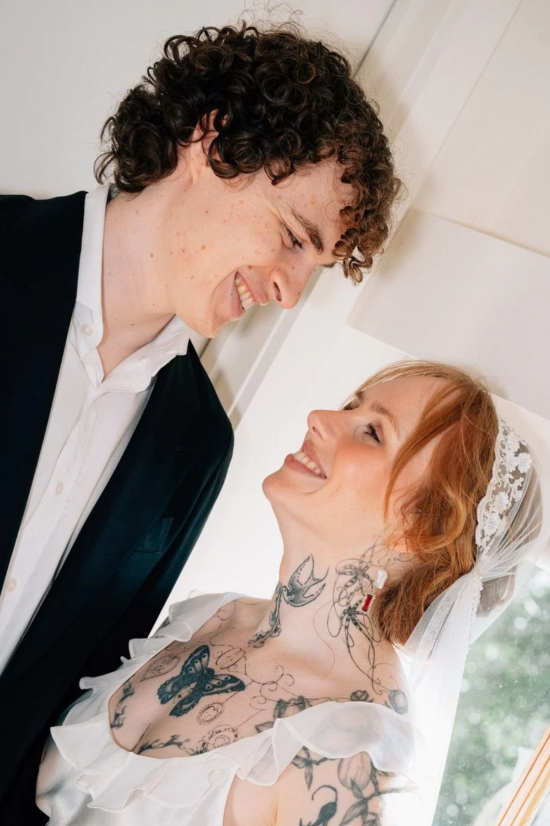 A bride and groom smiling at each other indoors, with the groom wearing a black suit and white shirt, and the bride having red hair, tattoos on her chest and neck, wearing a white dress and a lace veil.