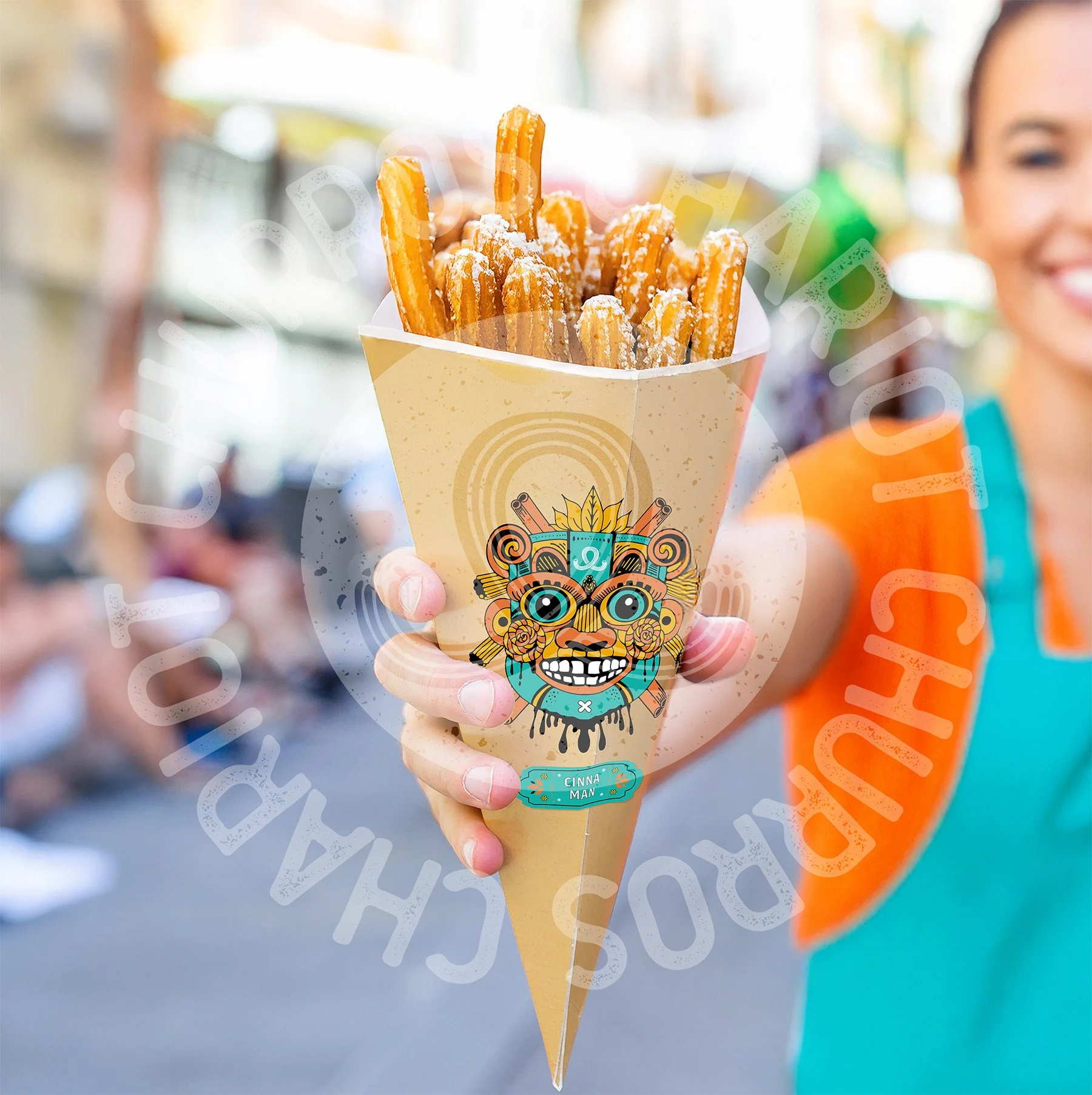 Person holding a cone of churros served in a decorative paper wrapper with a colorful face design, outdoors on a sunny day.