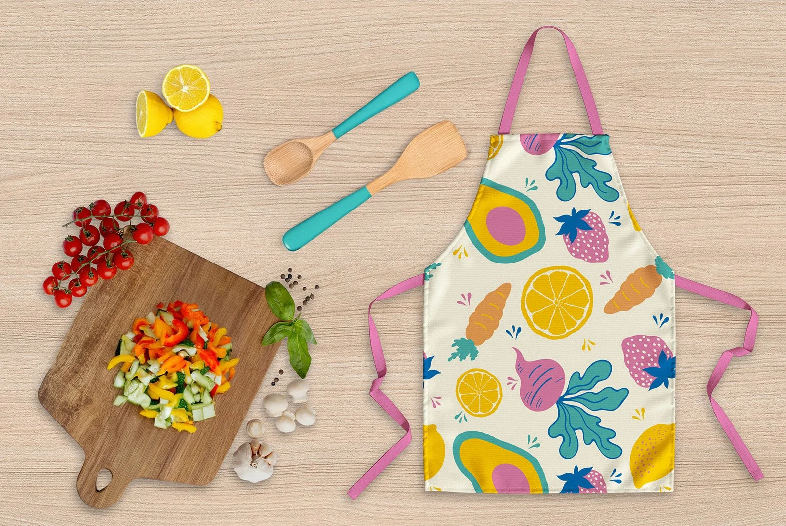 Kitchen apron with colorful fruit and vegetable pattern, wooden cutting board with chopped vegetables, cherry tomatoes, lemon halves, garlic, basil, and black peppercorns on a wooden surface, and two wooden cooking spoons.