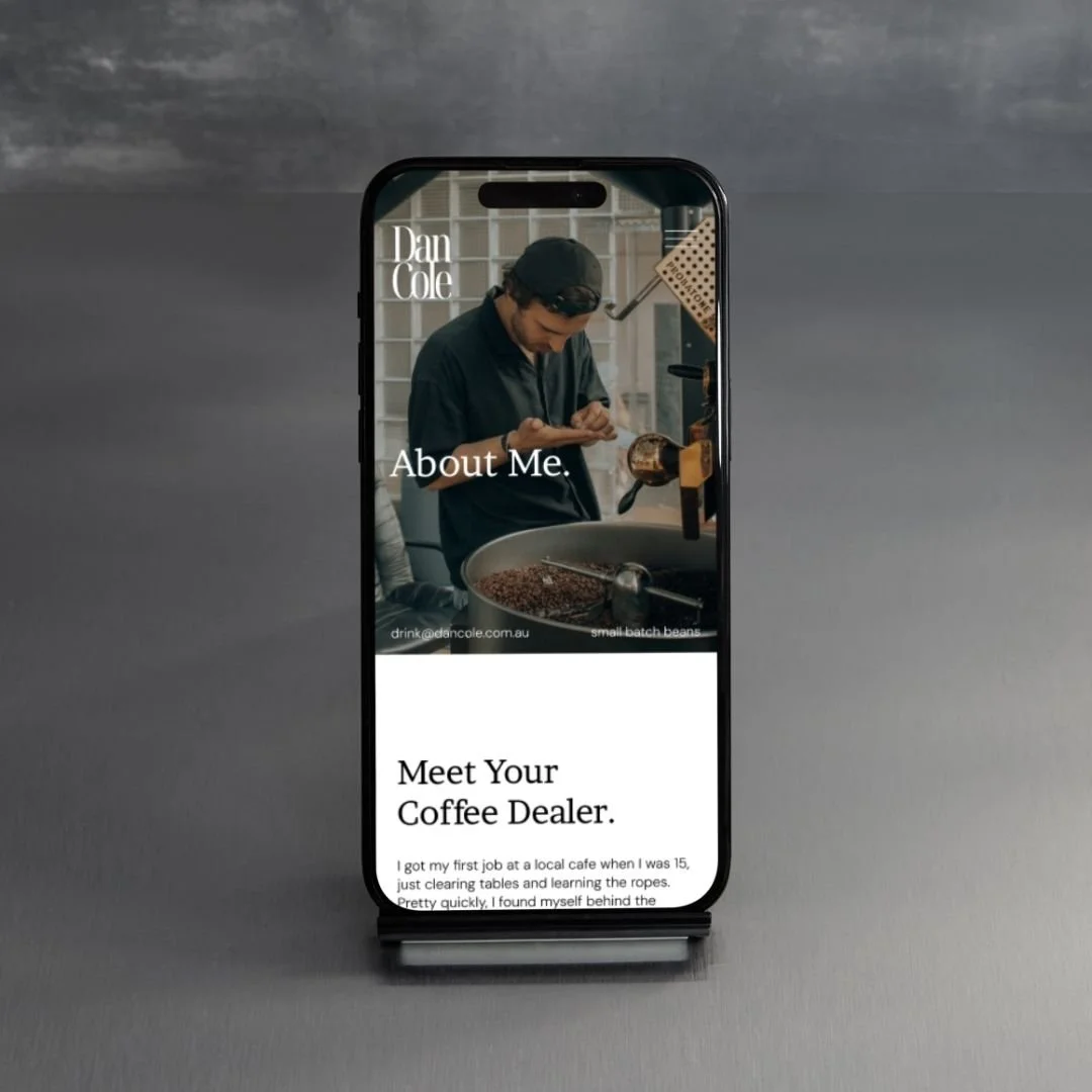 A smartphone displaying a website about a coffee dealer, with a photo of a man in a black shirt and black cap preparing coffee beans, on a gray background.