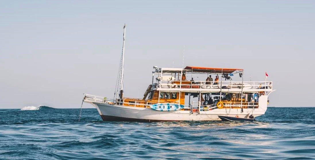 A wooden two-story boat with people on deck, floating on a calm sea under a clear sky.