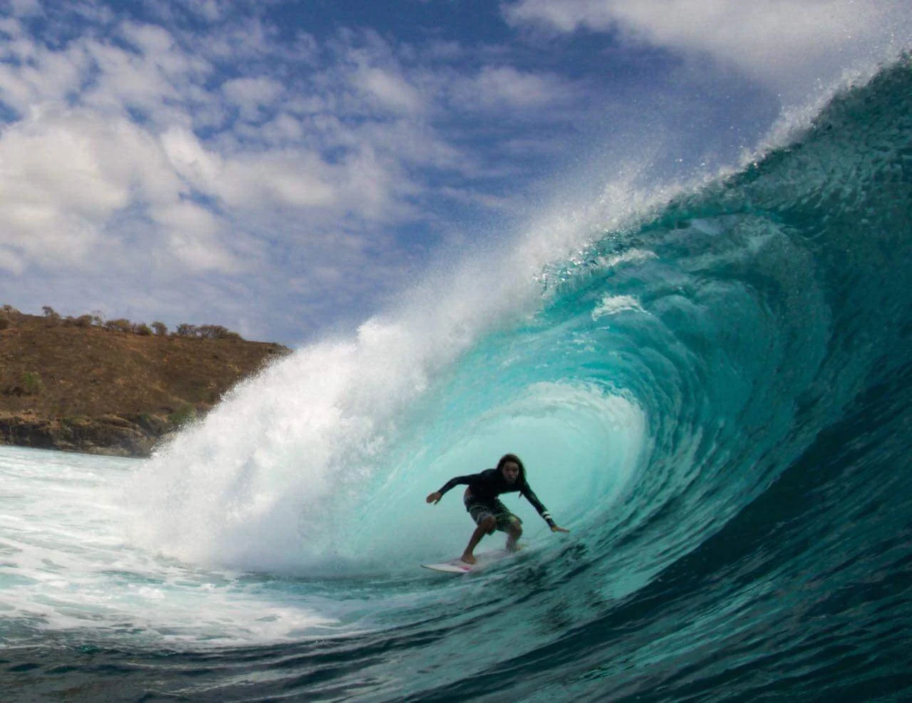 Surfer riding a wave tube with a clear blue sky and rugged coastline in the background.