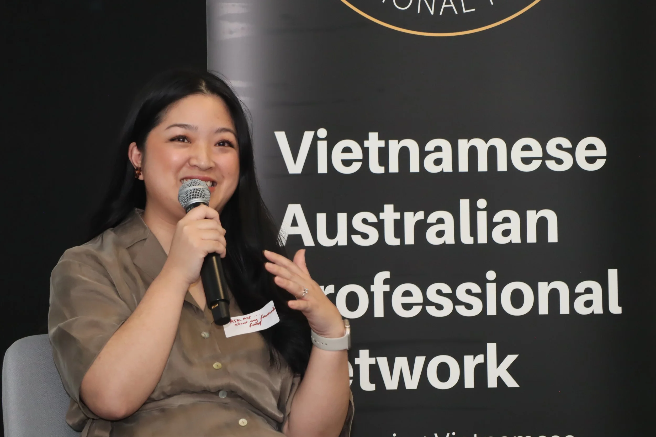 A woman with long black hair, wearing a light brown button-up shirt and a white watch, is speaking into a microphone while sitting on a gray chair. Behind her is a large black banner with white text that reads, 'Vietnamese Australian professional network.'