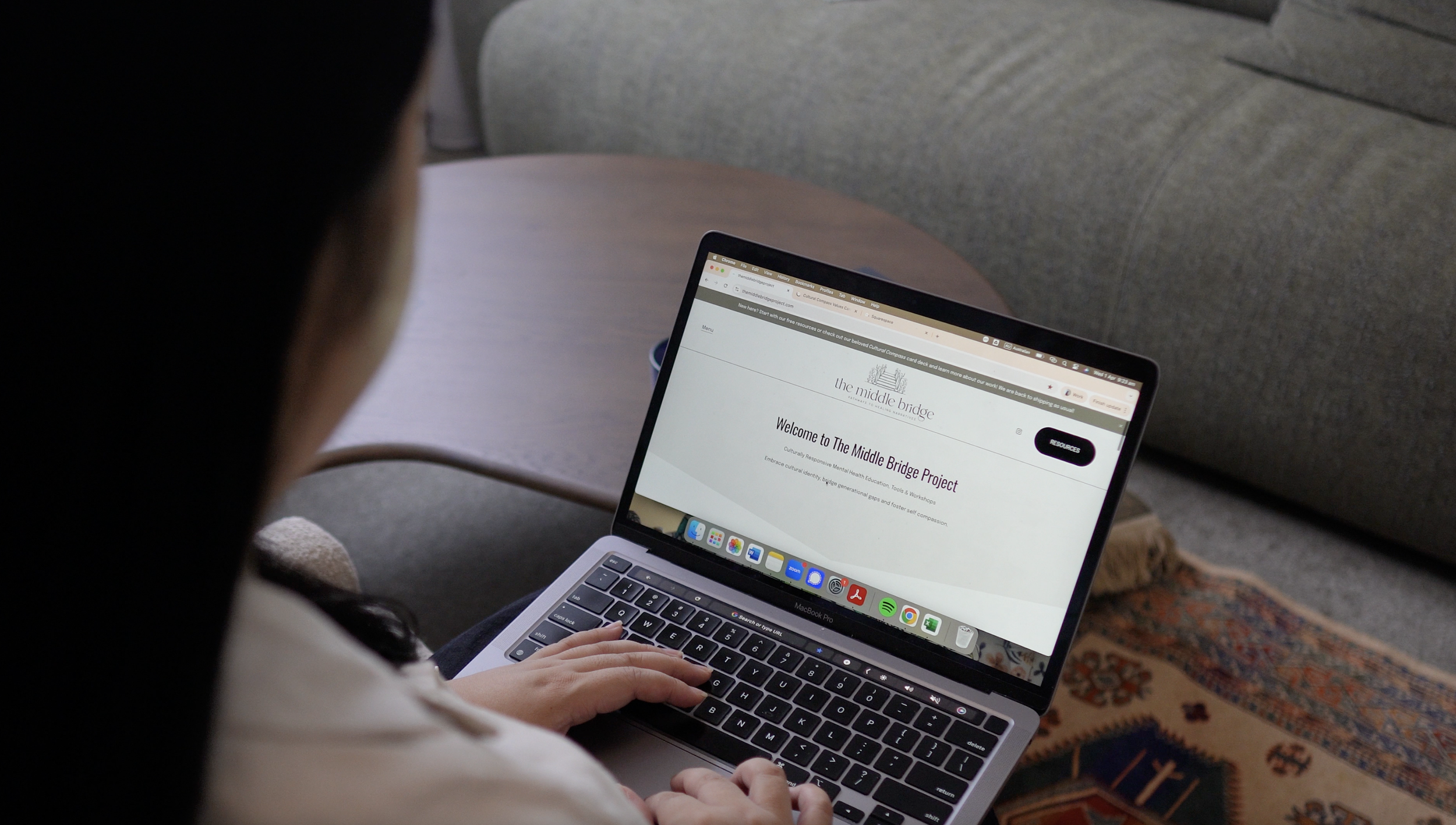 A person sitting on a patterned rug, using a silver laptop; the laptop screen displays a webpage titled "The Middle Bridge" with a message welcoming users to the project.