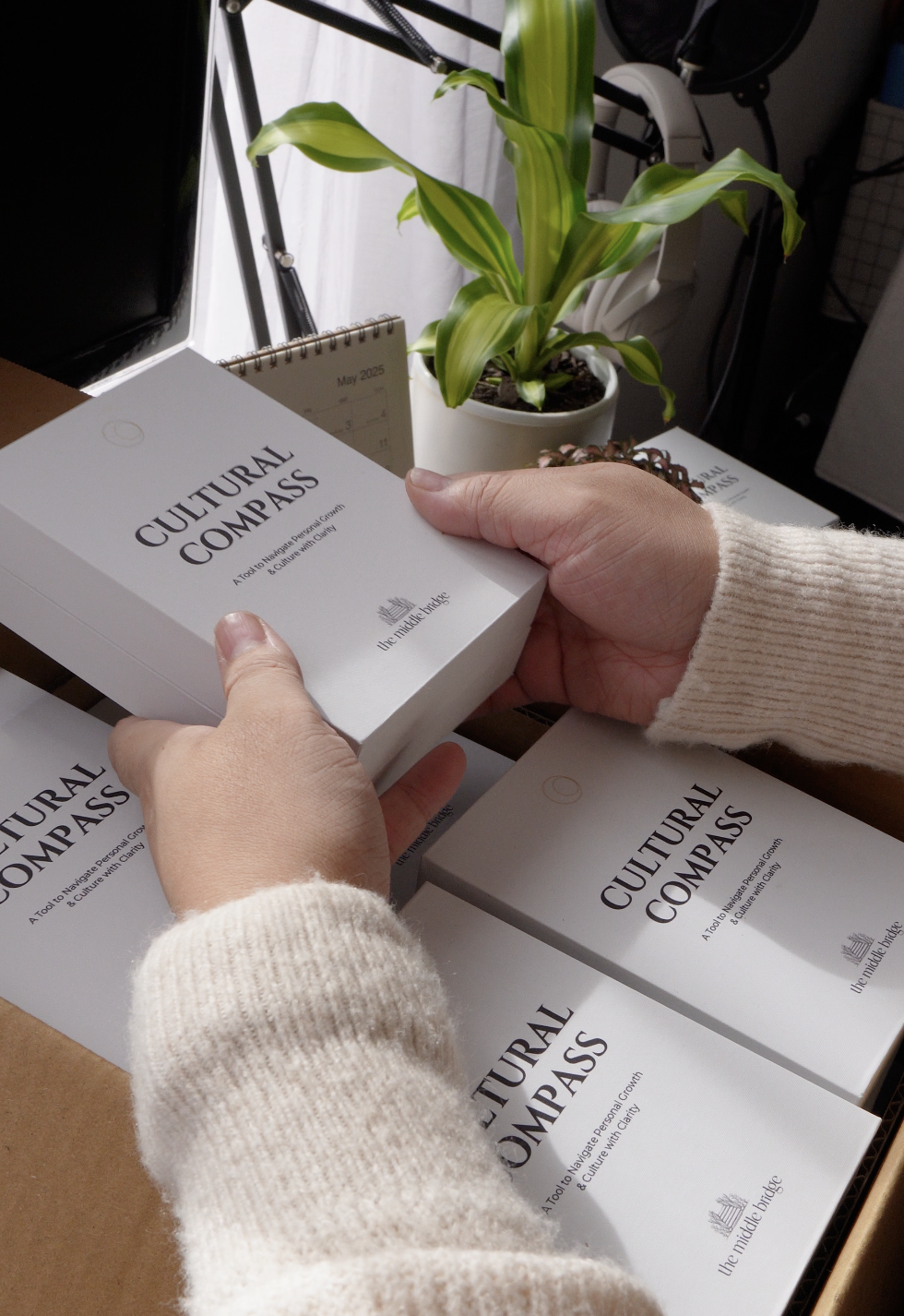 Person holding a book titled 'CULTURAL COMPASS' among other copies of the same book, with a green potted plant and a calendar on a desk in the background.