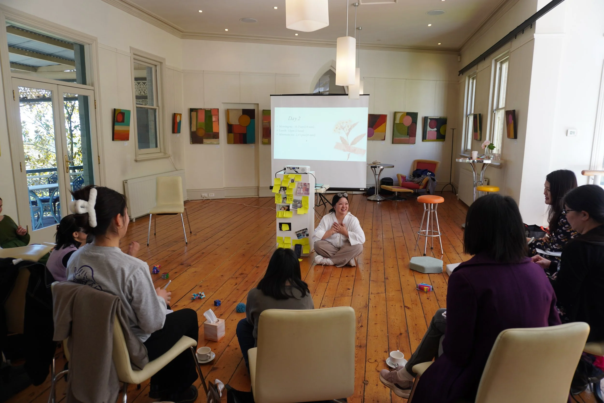 A group of people of colour in a circle in a bright room with wooden floors, listening to a woman who is sitting on the floor, smiling. There is a projection screen behind her with presentation slides, and colorful abstract artwork on the walls.