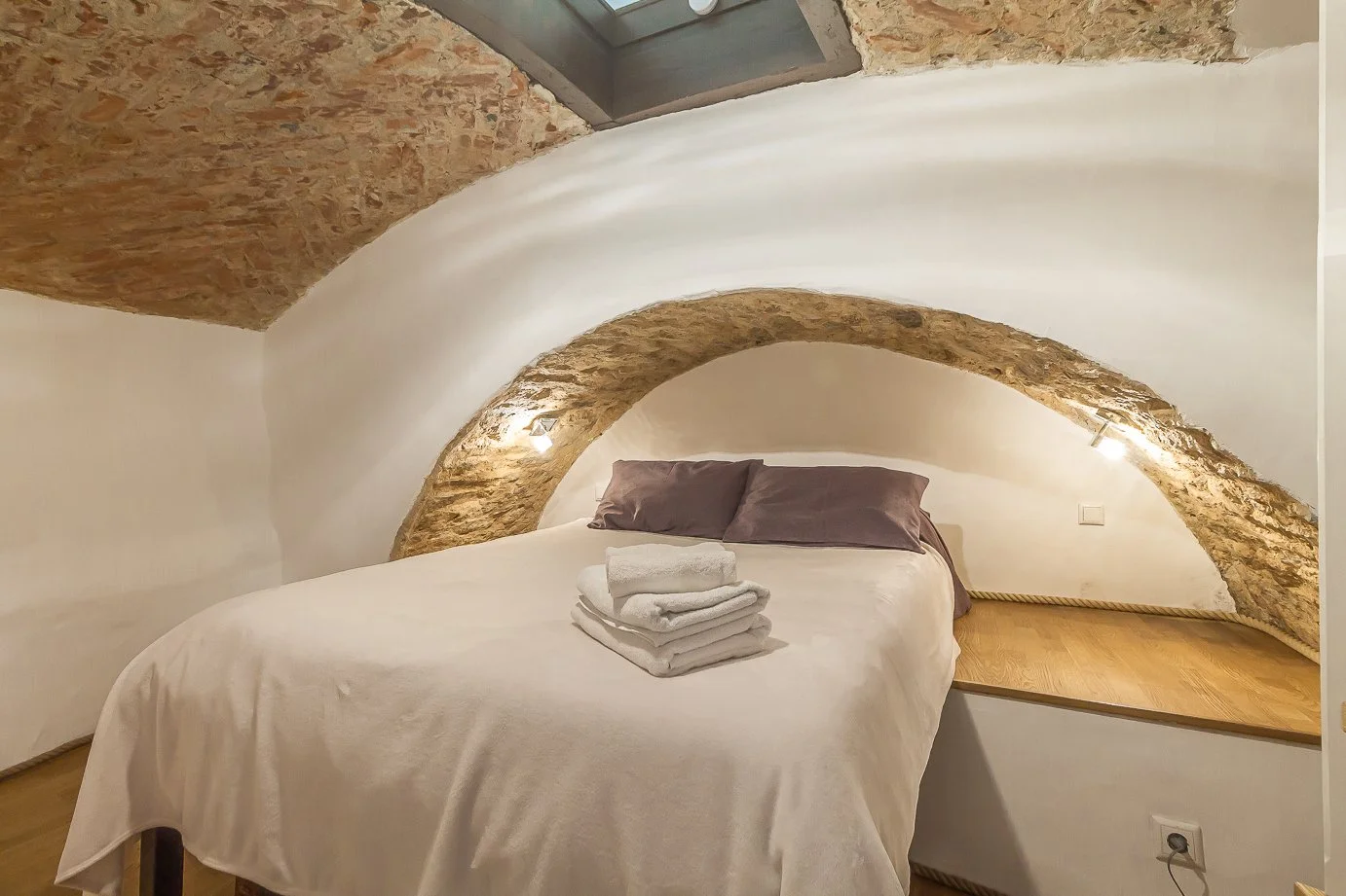A cozy bedroom with a bed under a stone arch in a rustic setting, with folded towels on the bed and soft lighting.