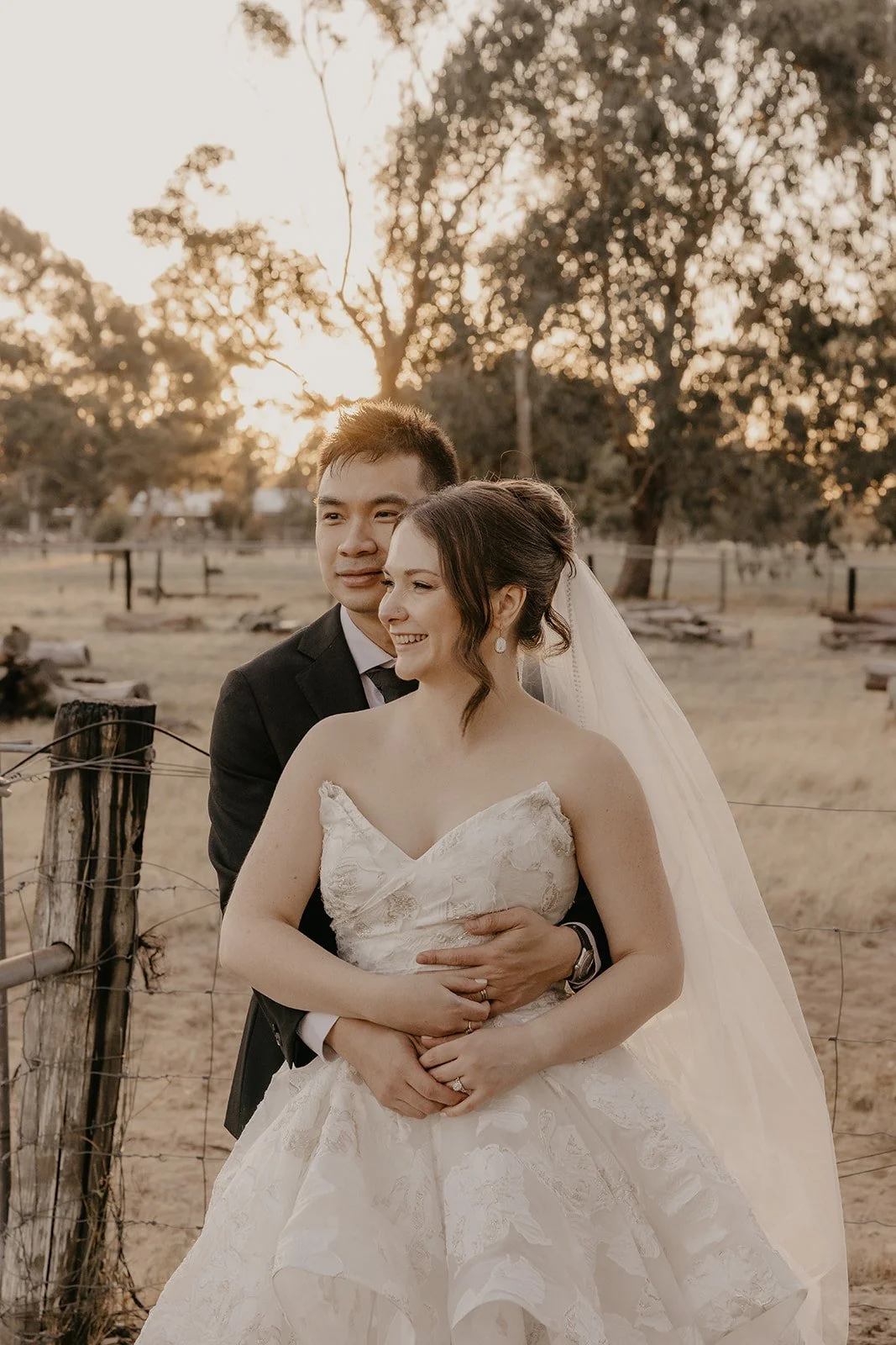 A newlywed couple in wedding attire standing outdoors in a field during sunset, with trees in the background. The groom is holding the bride from behind, both smiling.