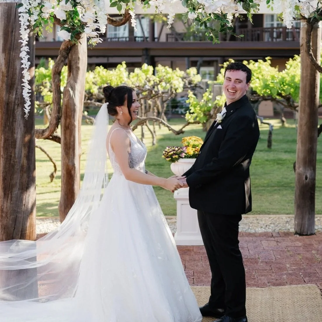 A bride and groom holding hands and smiling during their outdoor wedding ceremony under a floral arch.