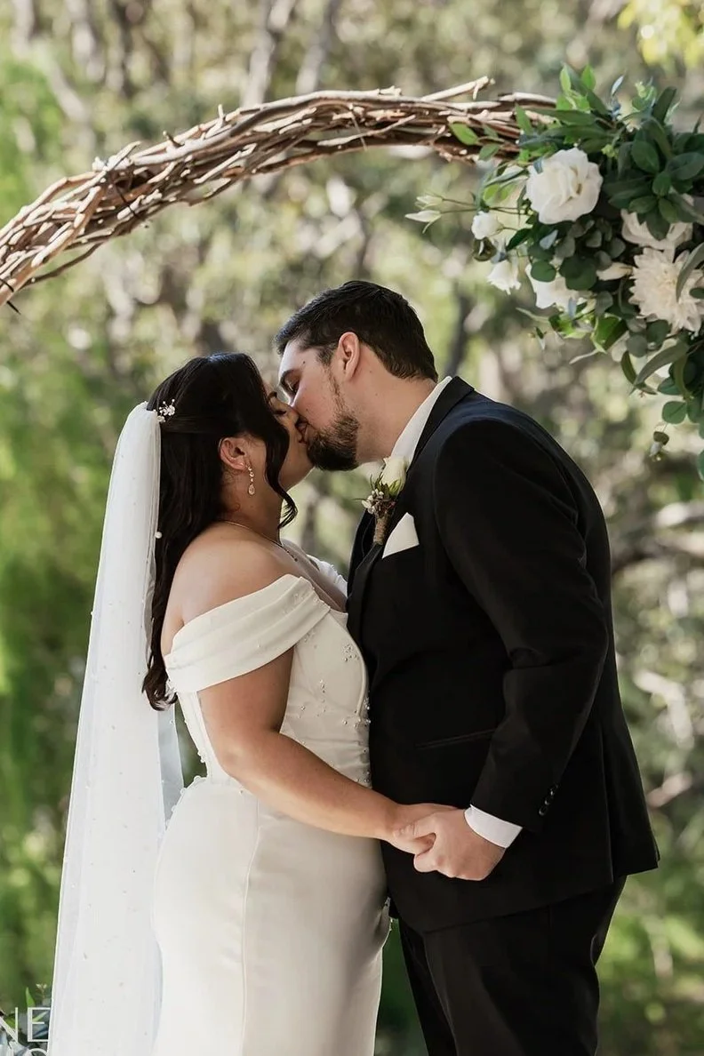 A bride and groom share a kiss during their wedding ceremony under a decorated arch with greenery and white flowers, set outdoors with blurred trees in the background.