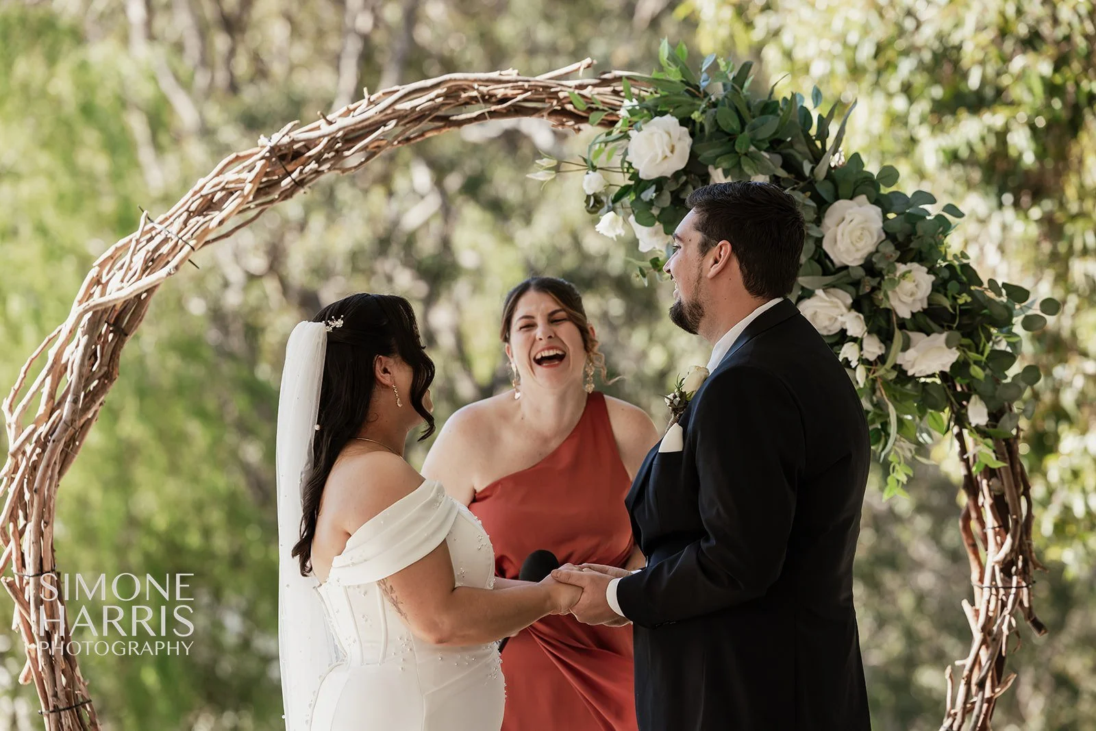 A bride and groom holding hands and laughing during their wedding ceremony outdoors under a floral arch, with an officiant standing between them.