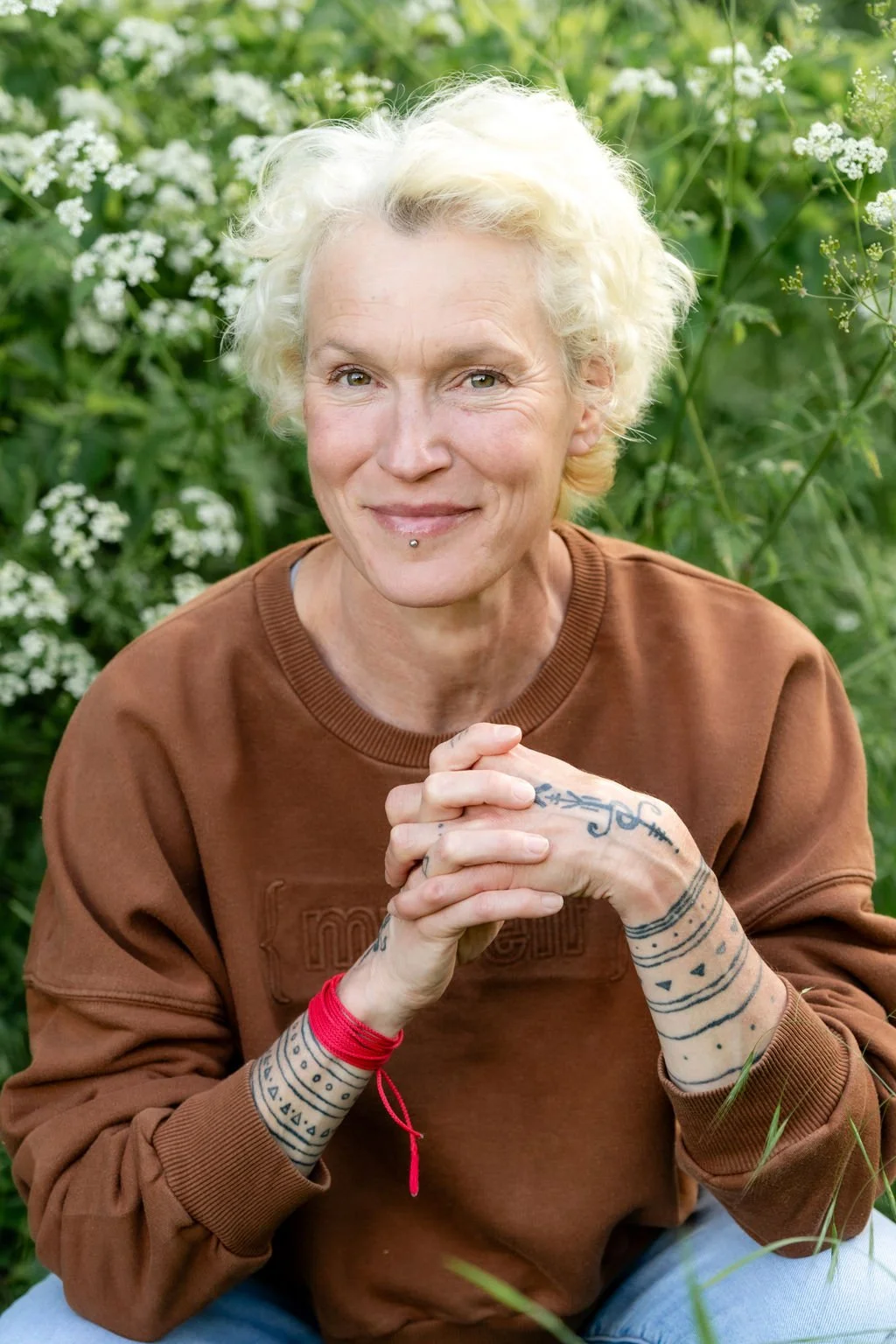 Kirsty Groves with short, curly, platinum blonde hair smiling and looking at the camera, sitting outdoors surrounded by green plants and white flowers, with her hands clasped.