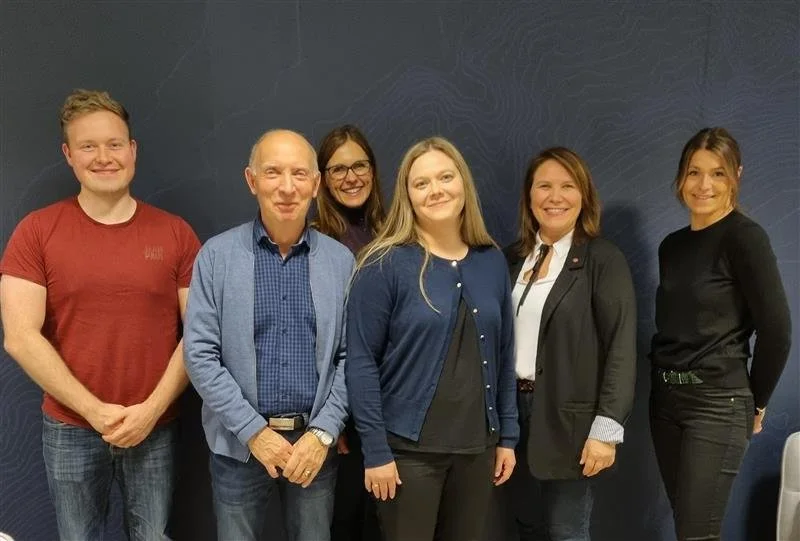 Sex mennesker som står foran en mørk blå vegg, smilende og poserer for fotografiet.