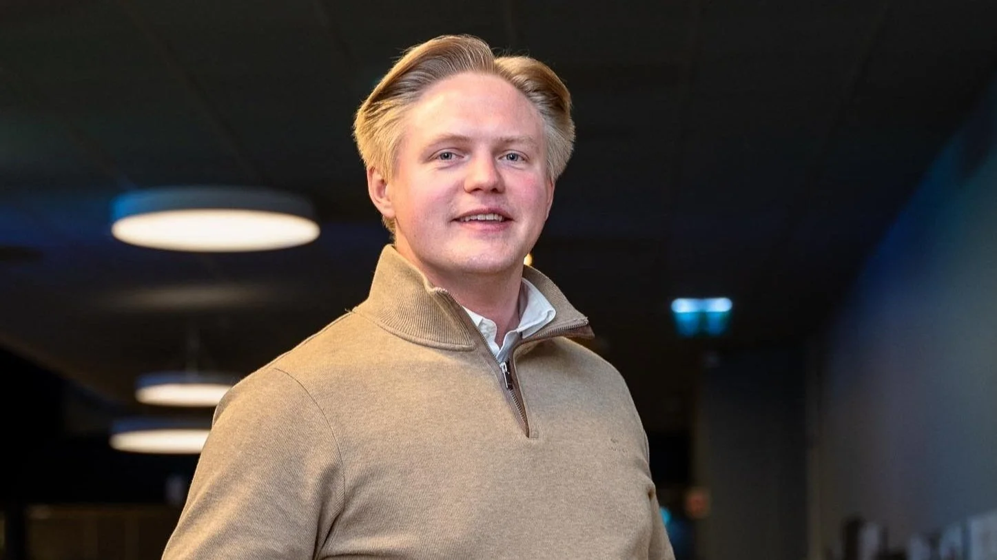 En ung mann med blond hår smilende, på mørk bakgrunn med taklamper.