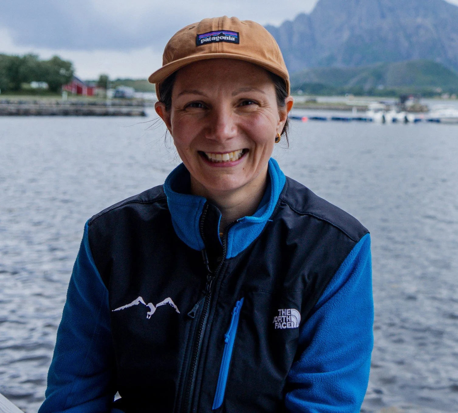 En kvinne smiler ved fjorden med fjell i bakgrunnen, iført en brun caps og en blå og svart jakke.