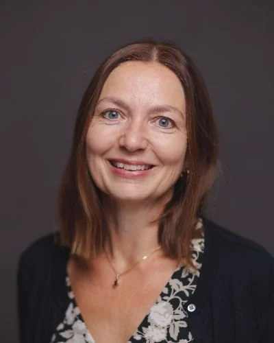 Portrett av en smilende kvinne med brunt hår, iført mørk blazer og blomstrete topp mot en grå bakgrunn.